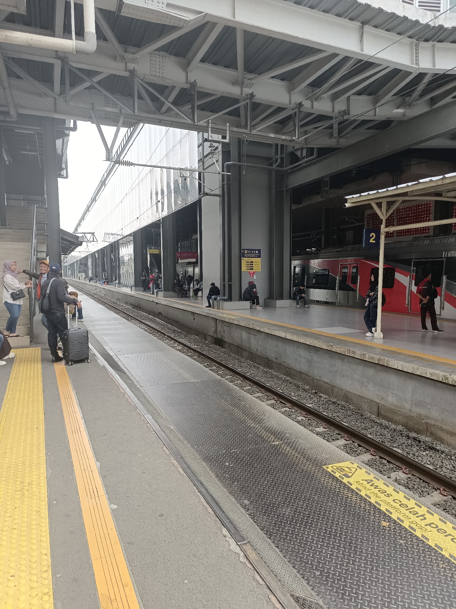 This image depicts a train station platform in Central Jakarta, Indonesia. The platform is elevated and constructed with concrete. Yellow tactile paving is visible along the edge of the platform, indicating a safety feature for visually impaired individuals. A metal railing runs along the edge of the platform, separating it from the train tracks. The tracks themselves are made of steel rails embedded in ballast.
Several individuals are present on the platform. In the foreground, a person stands with a dark suitcase on wheels. Further down the platform, a group of people are seated, awaiting a train. Some are looking at their phones, while others are engaged in conversation. A red and white train is visible parked at the platform, with its doors open. The train appears to be modern, with a sleek design.
The station architecture is characterized by a large, covered roof supported by steel beams and columns. The roof is made of corrugated metal. The background shows a multi-story building with a facade of reflective glass, suggesting it is part of a larger complex. Signage in Indonesian is visible on the platform, including a sign indicating "2" which likely refers to the platform number or train car. Another sign in yellow has text that translates to "Mind the platform gap." The overall impression is one of a busy, functional, and modern transportation hub.