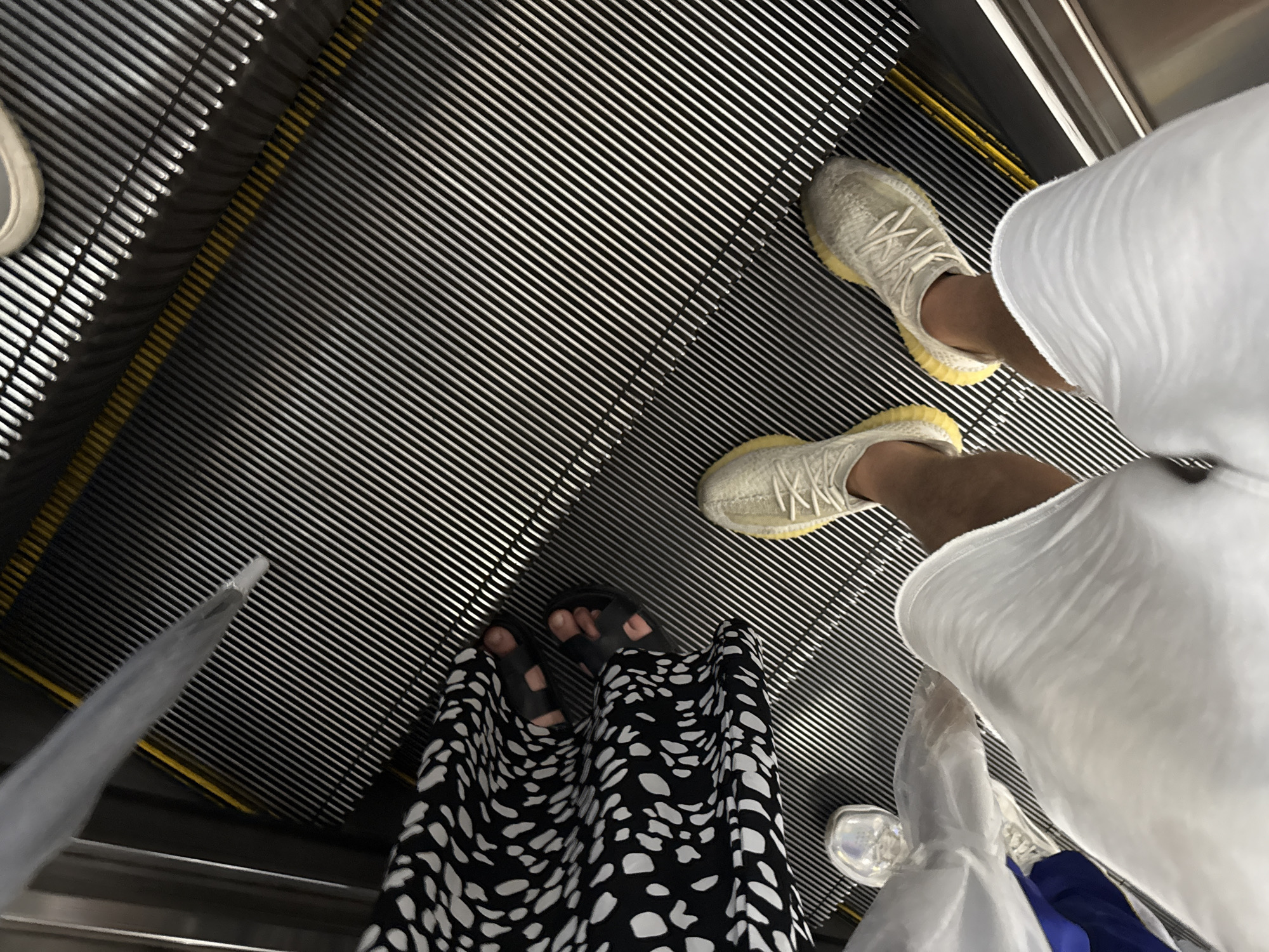 An overhead shot captures the lower legs and feet of two individuals on an escalator in the Haizhu District of China. The escalator's metal steps are black with thin yellow safety lines. One person wears a black and white patterned dress and black sandals. The other person wears white shorts and light-colored athletic shoes with yellow soles. Another person's foot, wearing a silver shoe, is visible at the bottom left. The scene appears to be an indoor public space, possibly a mall or transportation hub.