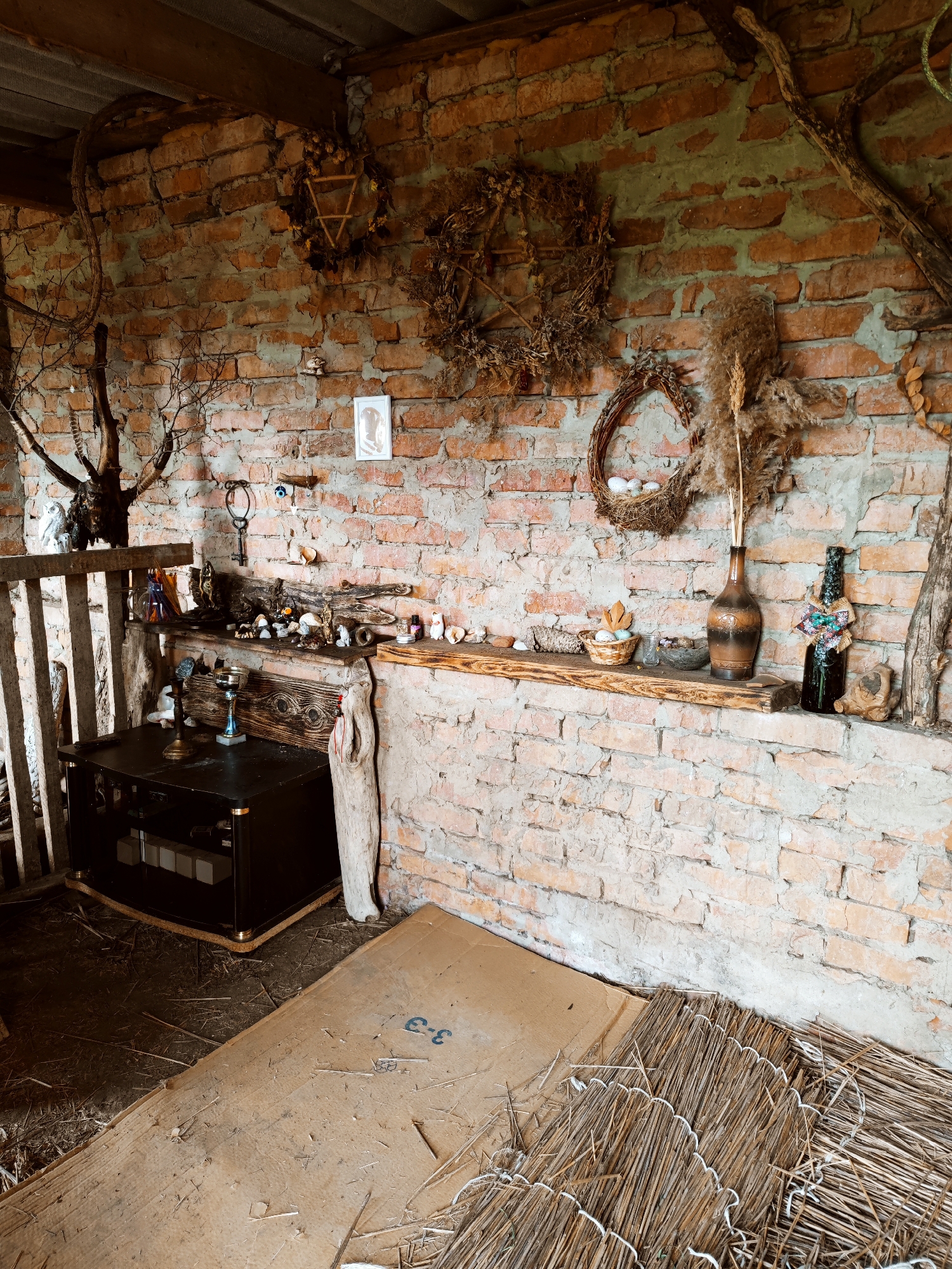 The image depicts the interior of a room with brick walls. Wreaths and dried plants are hanging on the wall, along with a framed picture and other decorative items. Two wooden shelves display various objects including vases, figurines, and other small items. Below the shelves, there is a black cabinet and a piece of cardboard lying on the floor, amidst some straw-like material. The ceiling appears to be corrugated metal. There is no indication of the city of Yubileyny, Russia, in the image.