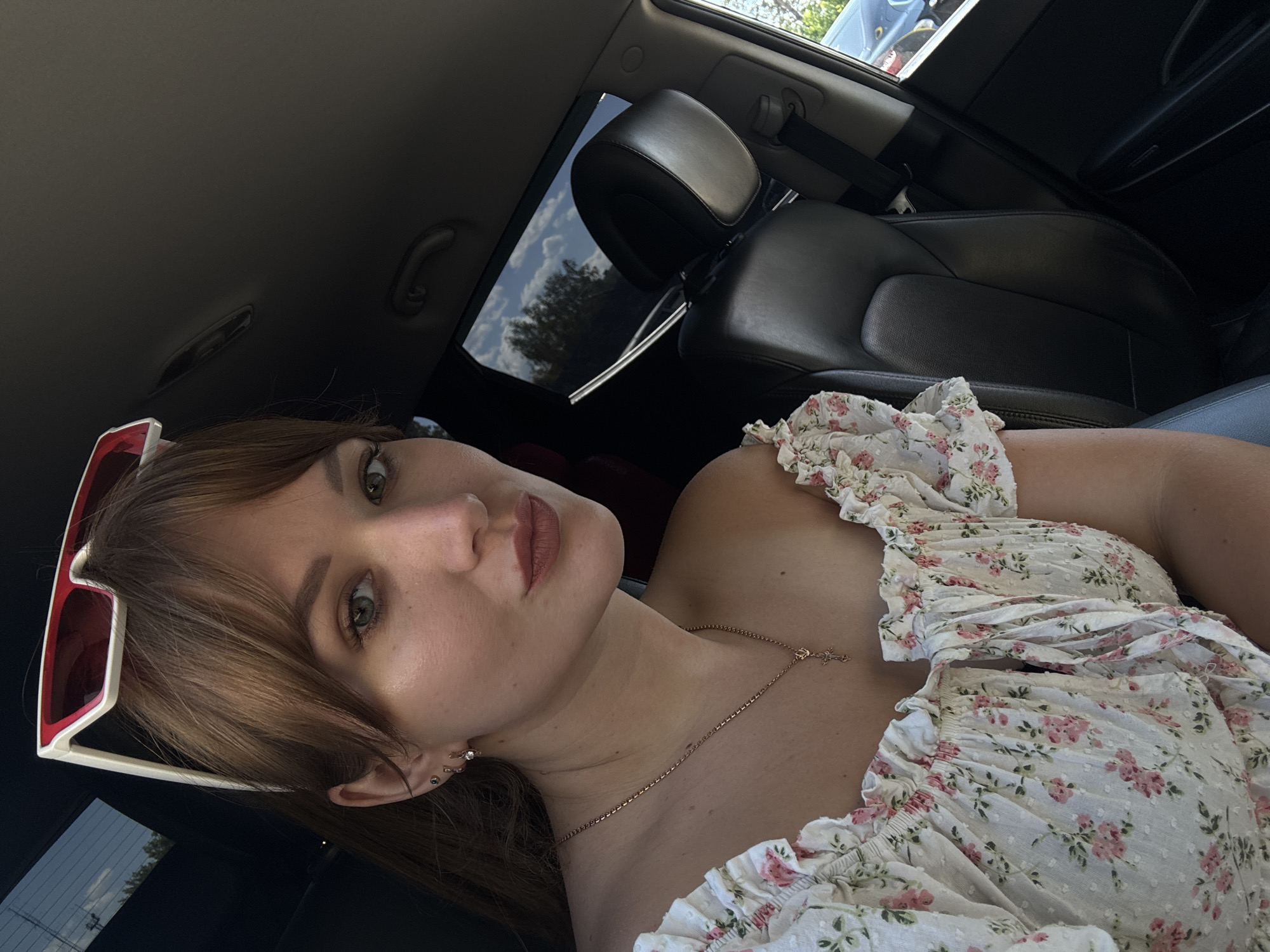 In Chaikovsky, Russia, a woman is captured in a selfie taken inside a car. She has light skin, green eyes, and shoulder-length brown hair with bangs. She is wearing a white off-the-shoulder top with a floral pattern of pink roses and green leaves. A delicate gold necklace with a cross pendant rests on her collarbone, and she has two small earrings in her left ear. White-rimmed sunglasses with red lenses are perched on top of her head. The interior of the car is visible, including a black leather seat and a portion of the car's roof. Through the tinted rear window, trees and a cloudy blue sky are discernible, indicating an outdoor setting. The overall scene is casual and suggests a daytime outing.