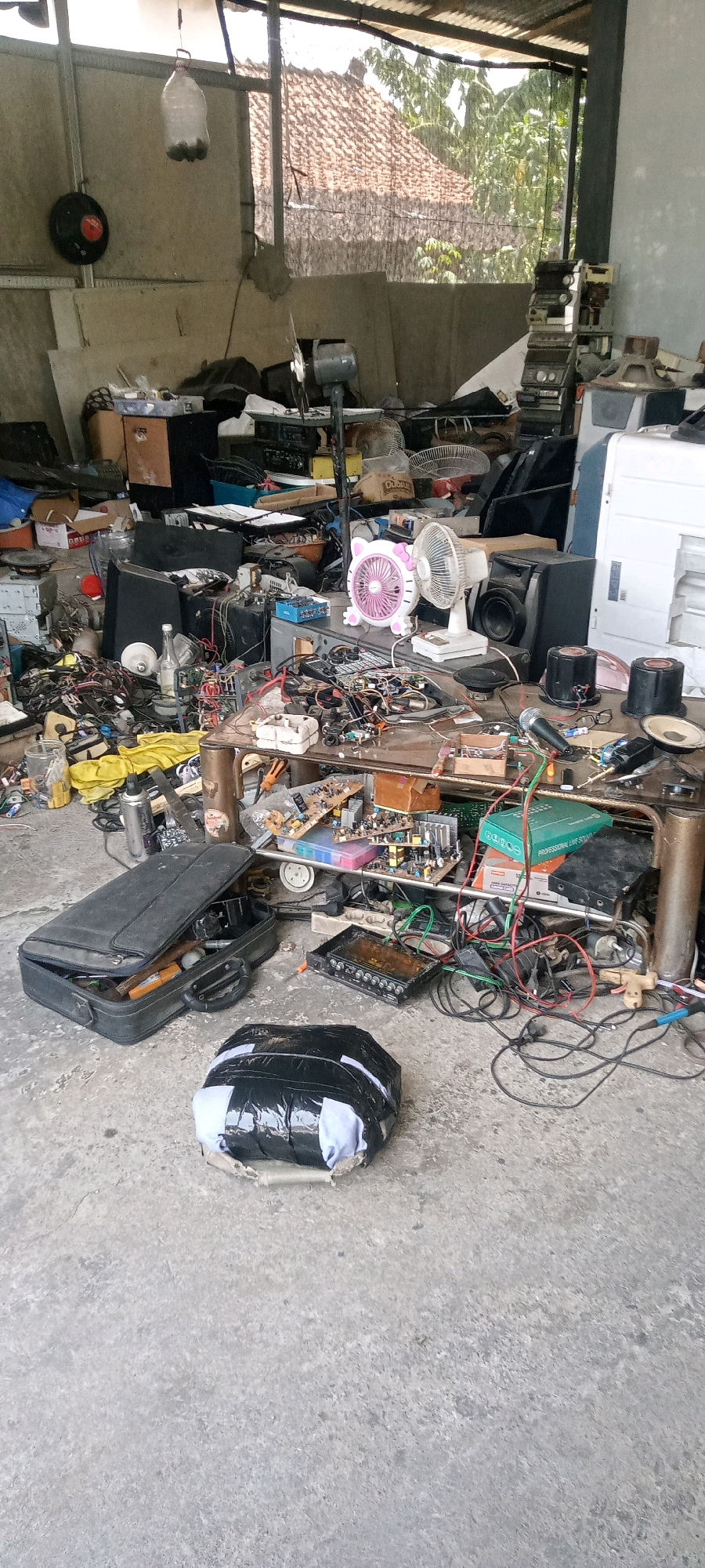 The image depicts a cluttered, open-air workshop or storage area, likely located in Malangan, Indonesia. The scene is characterized by a large accumulation of electronic equipment, spare parts, tools, and various discarded items. In the foreground, a black, rectangular bag is wrapped in black plastic and secured with tape, with a light-colored fabric peeking out. To the left of this bag is an open black briefcase containing various objects.
Dominating the mid-ground is a table laden with electronic components, circuit boards, wires, fans, and speakers. A pink and white Hello Kitty-themed fan is prominently displayed alongside a white desk fan. Various boxes, some made of cardboard and others of plastic, are stacked and scattered throughout the space. Electrical wires are tangled across the floor and table, suggesting a chaotic working environment. In the background, a large speaker cabinet and other electronic equipment are visible, along with portions of a corrugated metal roof and tiled buildings. The overall impression is one of disarray and potential salvage or repair work involving old electronics. There are no people visible in the image.