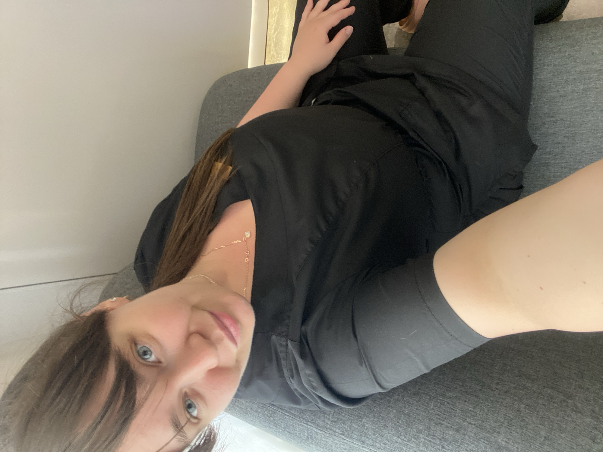 A woman with light skin, blue eyes, and brown hair is lying on a grey upholstered surface, possibly a couch or a bench. She is wearing black clothing, likely scrubs, and a delicate gold necklace. Her body is angled diagonally across the frame, with her head tilted towards the lower-left corner. The background is a light grey or white wall. The location is not apparent from the image and cannot be determined to be Votkinsk, Russia.