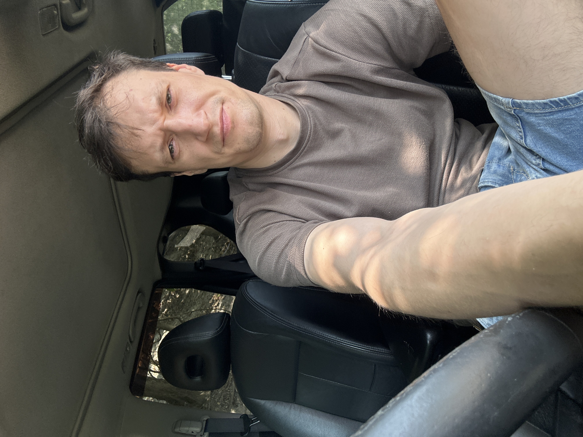 A man sits in the driver's seat of a car, looking at the camera with a slight frown. He is wearing a brown t-shirt and denim shorts. The car's interior is visible, including the black leather seats, steering wheel, and sunroof. The scene appears to be outdoors, with trees and foliage visible through the car windows. The lighting suggests it is daytime. The location is not specified in the image.