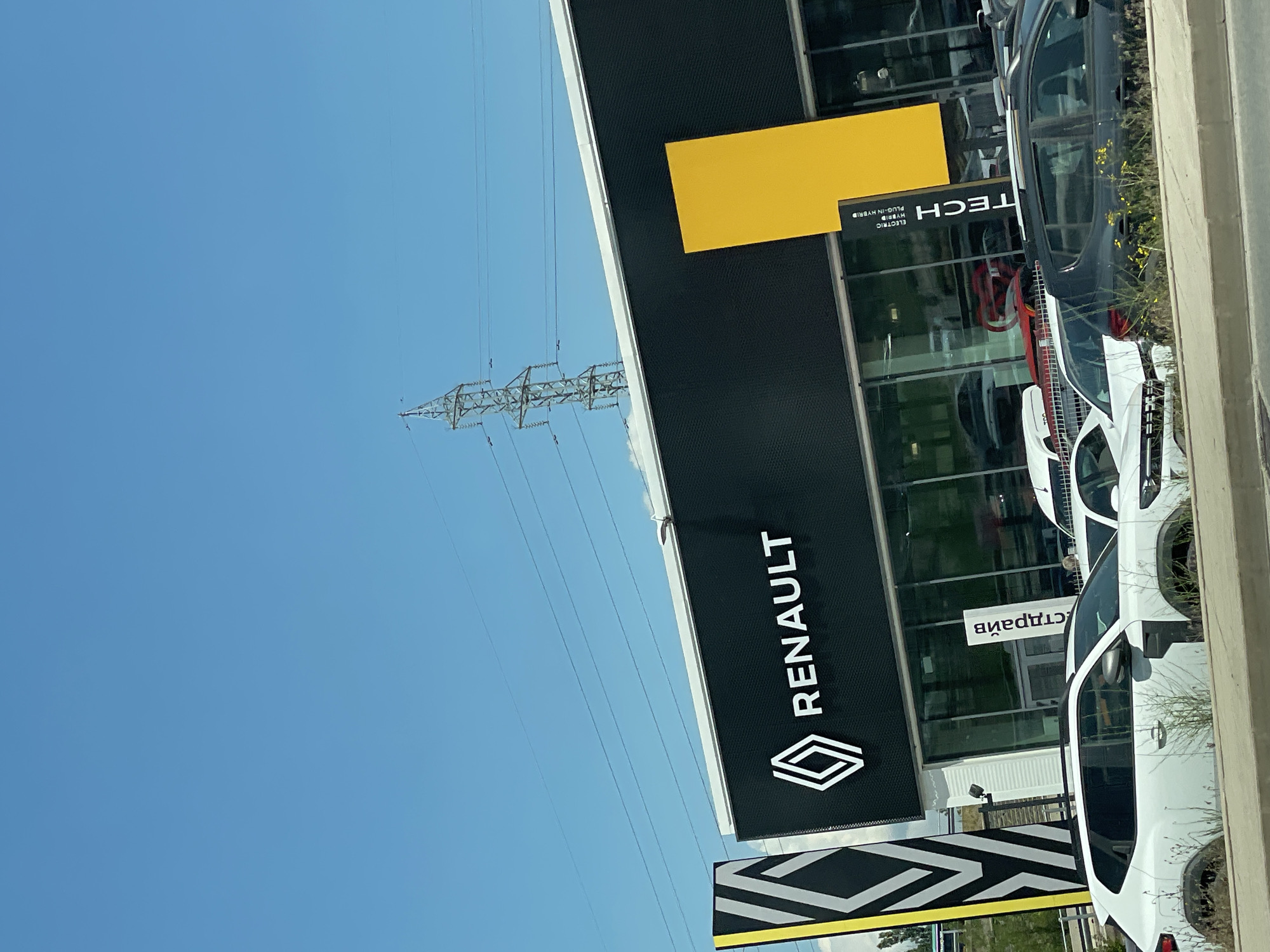 The image shows the exterior of a Renault dealership in Sofia, Bulgaria. The building has a black facade with the Renault logo and name prominently displayed. A large yellow rectangular sign is mounted on the building. In front of the dealership, several cars are parked, including white SUVs and a dark-colored hatchback. The sky is clear blue with power lines and a transmission tower visible in the background. The lighting suggests it is daytime.