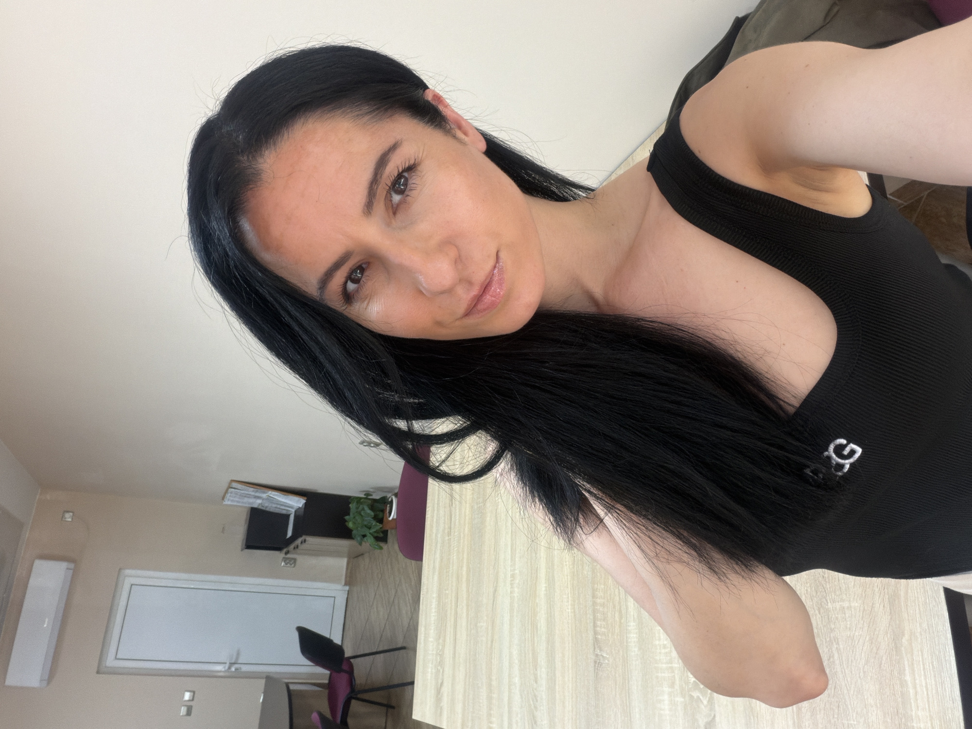 A medium shot captures a woman indoors, with a glimpse of a room in the background. She is positioned at an angle, looking toward the camera with a slight smile. Her dark hair falls to one side, contrasting with her complexion. She wears a black top with white letters near the shoulder.
The background reveals a partially visible interior space. A wooden-textured surface runs along the foreground. Behind it, a wall features a white window, an air conditioner, and a dark chair with a purple back. A plant sits atop a unit behind the chair.
While a specific location in Pleven, Bulgaria, cannot be identified from the details available in the image, the presence of an air conditioner unit and the particular interior design features suggest a residential or commercial setting within the city.