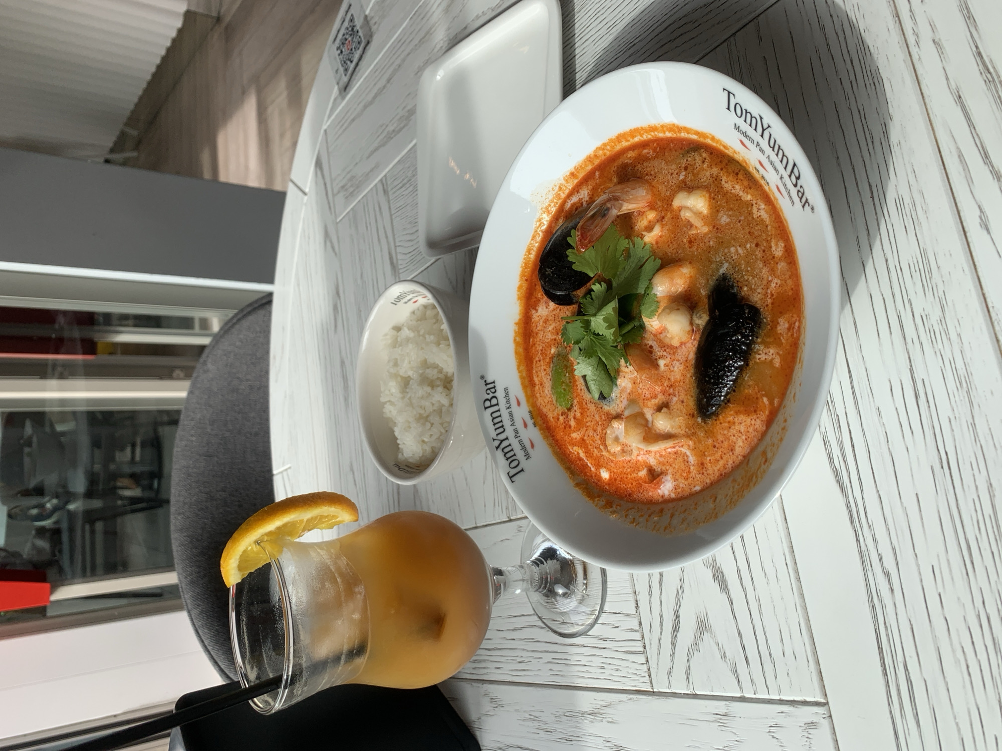 Here is a concise description of the image:
The image shows a meal from the restaurant "TomYum Bar," a Modern Pan-Asian Kitchen, located in Almaty, Kazakhstan. The meal consists of a bowl of Tom Yum soup garnished with herbs, served with a side of rice and a glass of orange-colored drink with a slice of orange on the rim. The food is presented on a white, herringbone-patterned table, likely in the restaurant. A chair is visible, suggesting an indoor setting.