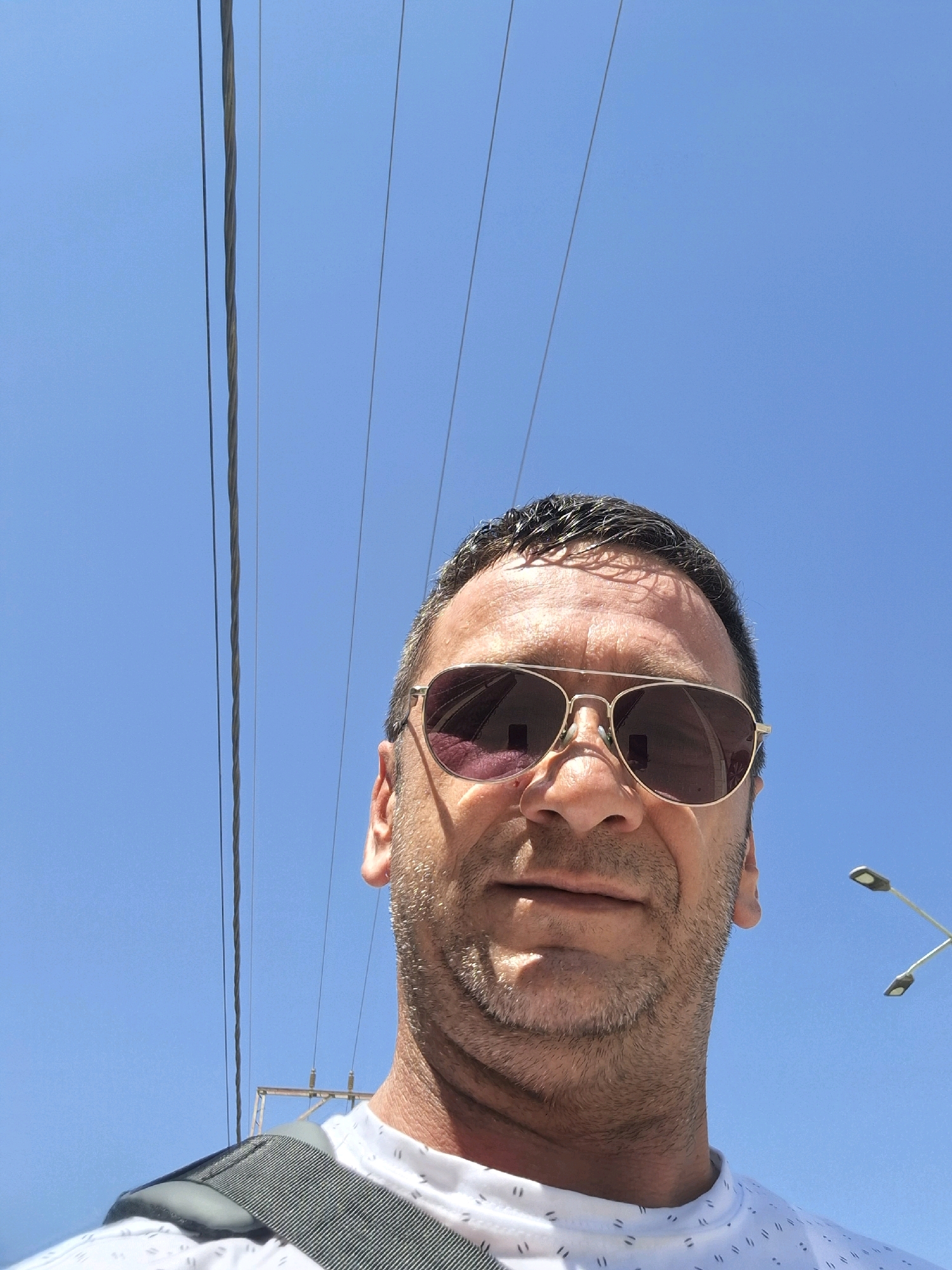 A close-up, low-angle selfie is taken outdoors against a clear blue sky in Dimona, Israel. The subject is a middle-aged man with short, dark hair, wearing aviator sunglasses with brown lenses and a white t-shirt with a subtle pattern. He has stubble on his face. In the background, several thick power lines stretch across the frame, with a utility pole visible on the right side. The sun casts shadows on the man's face, indicating it is daytime. The overall impression is that of a casual, outdoor snapshot.