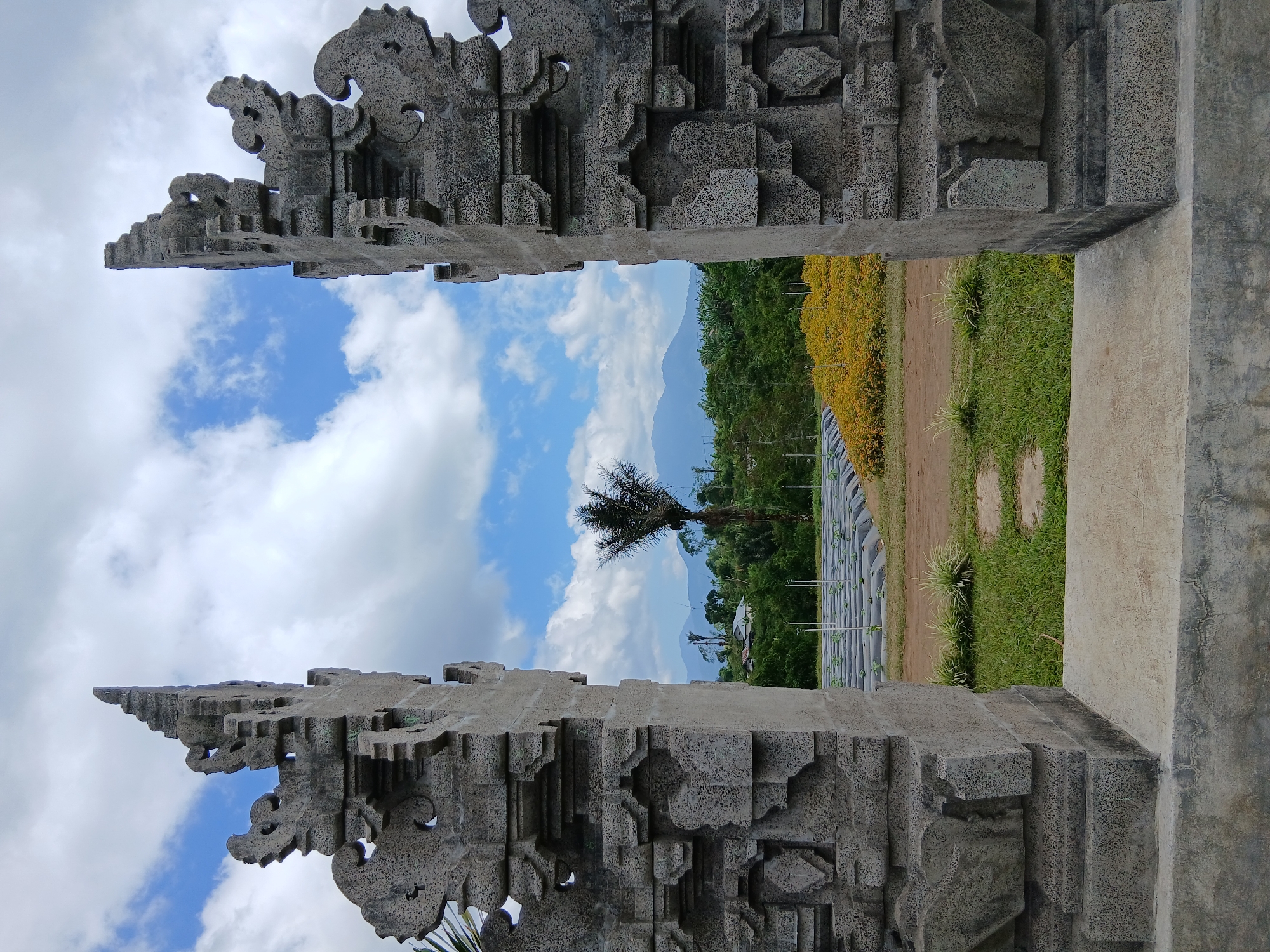 This is a full-frame, eye-level, outdoor shot captured on a bright, cloudy day. The image is framed by two intricately carved gray stone gate posts, possibly a Candi Bentar gate, commonly found in Bali, Indonesia. These posts, with their detailed traditional motifs, stand prominently in the foreground, framing the view beyond. Through the gateway, a lush landscape unfolds. In the mid-ground, a field of vibrant yellow flowers is visible, bordered by rows of what appear to be young plants covered with black plastic sheeting, suggesting an agricultural setting. A solitary palm tree stands tall near the center of the frame, its dark silhouette contrasting with the bright sky and green foliage. Beyond the fields and vegetation, a mountain range with soft blue peaks rises against a sky filled with white, fluffy clouds and patches of clear blue. The scene evokes a sense of serene natural beauty within a culturally rich environment, characteristic of the Kintamani region in Bali, known for its volcanic landscapes and traditional architecture. There are no people or animals visible in the image.