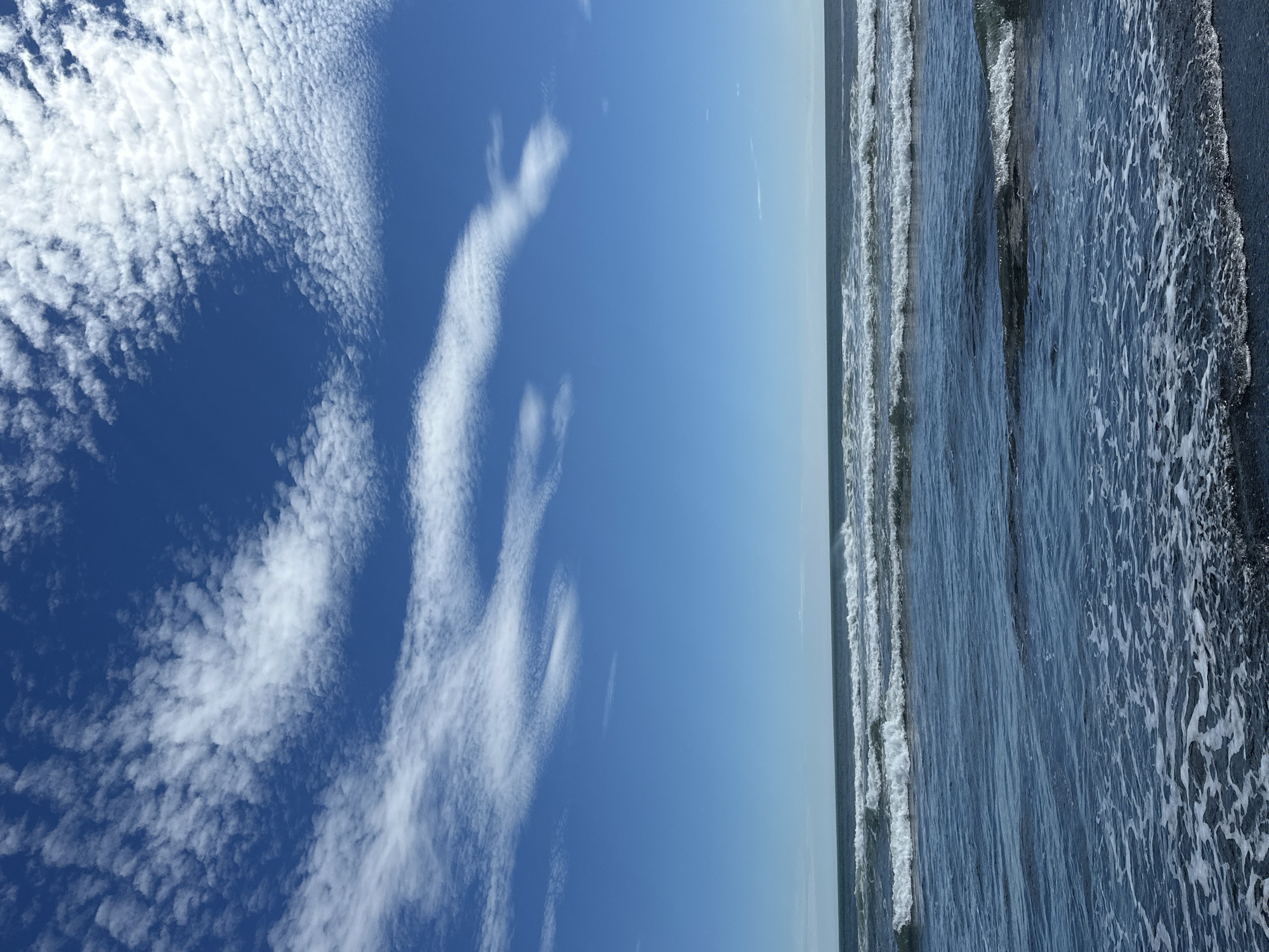 The image depicts a daytime scene at a beach. The sky is a vibrant blue, sparsely populated with scattered white, wispy clouds. The ocean stretches across the frame, with several rows of waves rolling towards the shore. The water is a deep blue, and white foam is visible where the waves break on the dark sand. The shoreline is visible in the foreground, with the dark sand wet and glistening from the receding water. There are no people or man-made structures visible in the image. The scene is peaceful and natural.
This image does not depict Petropavlovsk-Kamchatsky, Russia.