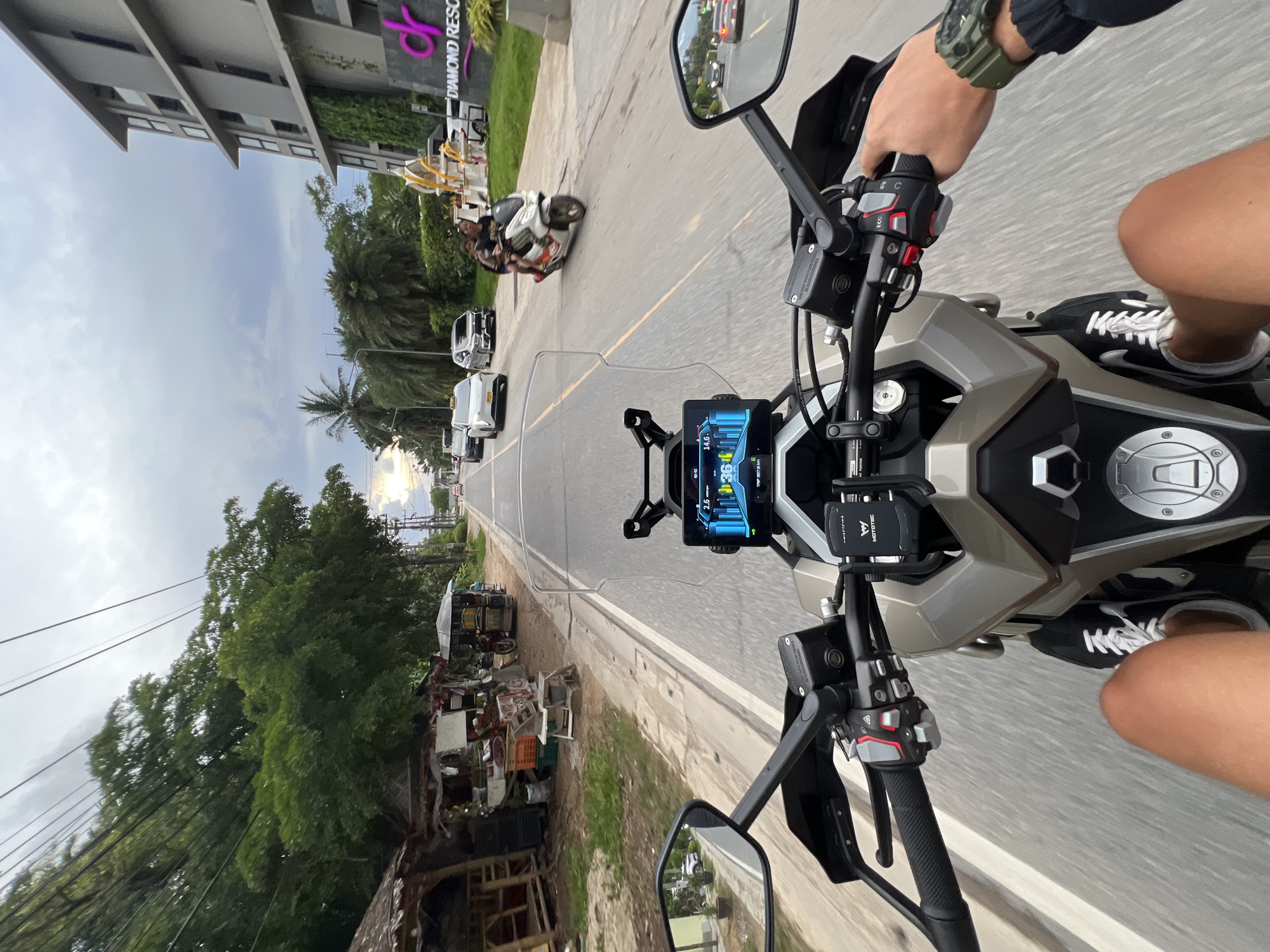 A first-person perspective shot shows a person riding a motorcycle down a paved road. The motorcycle has a digital display screen and a phone mount attached to the handlebars. The rider's legs and feet, wearing sneakers, are visible. The road is lined with trees, small shops, and a modern hotel building with the sign "DIAMOND RESORT." Several cars and a scooter with two passengers are also on the road. The sky is cloudy.
