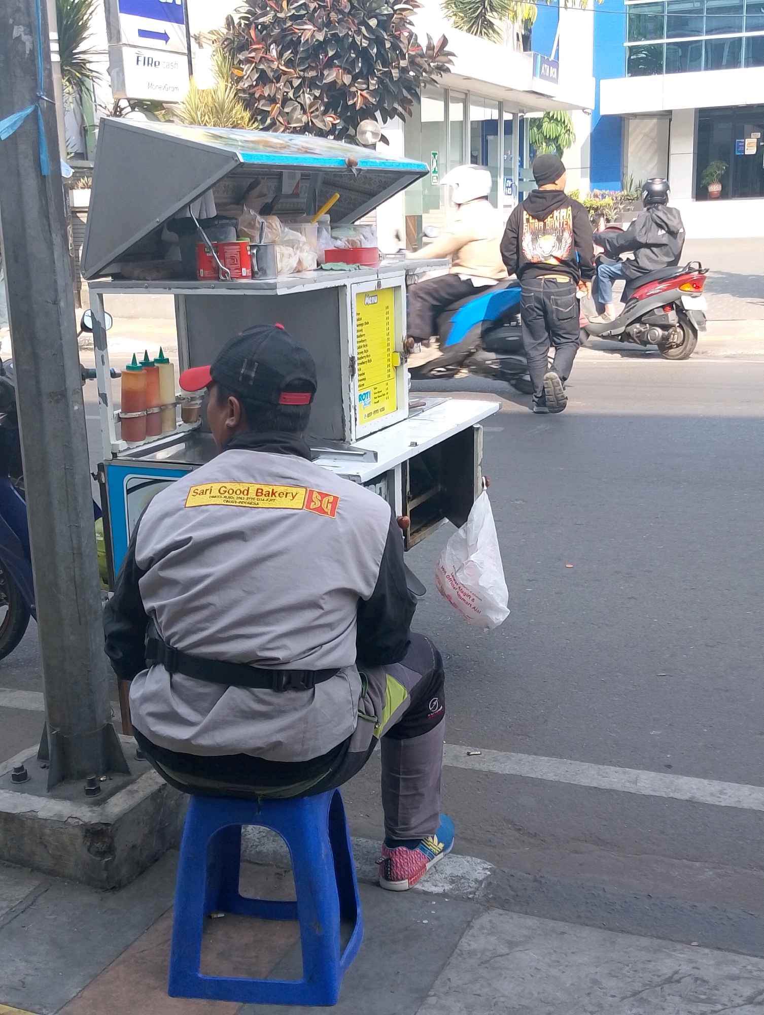 In Lembang, Indonesia, on a sunny day, a street food vendor sits on a blue plastic stool facing away from the camera, his back to a cart. He wears a grey and black jacket with "Sari Good Bakery SG" printed on the back in yellow. The cart has a metal canopy holding various food items and condiments. Behind him, two men on motorcycles ride past, one in a black hoodie and jeans, the other in a black hoodie with a helmet. The background shows a building with blue accents and lush greenery. The scene captures a typical street view with commercial activity and urban elements.