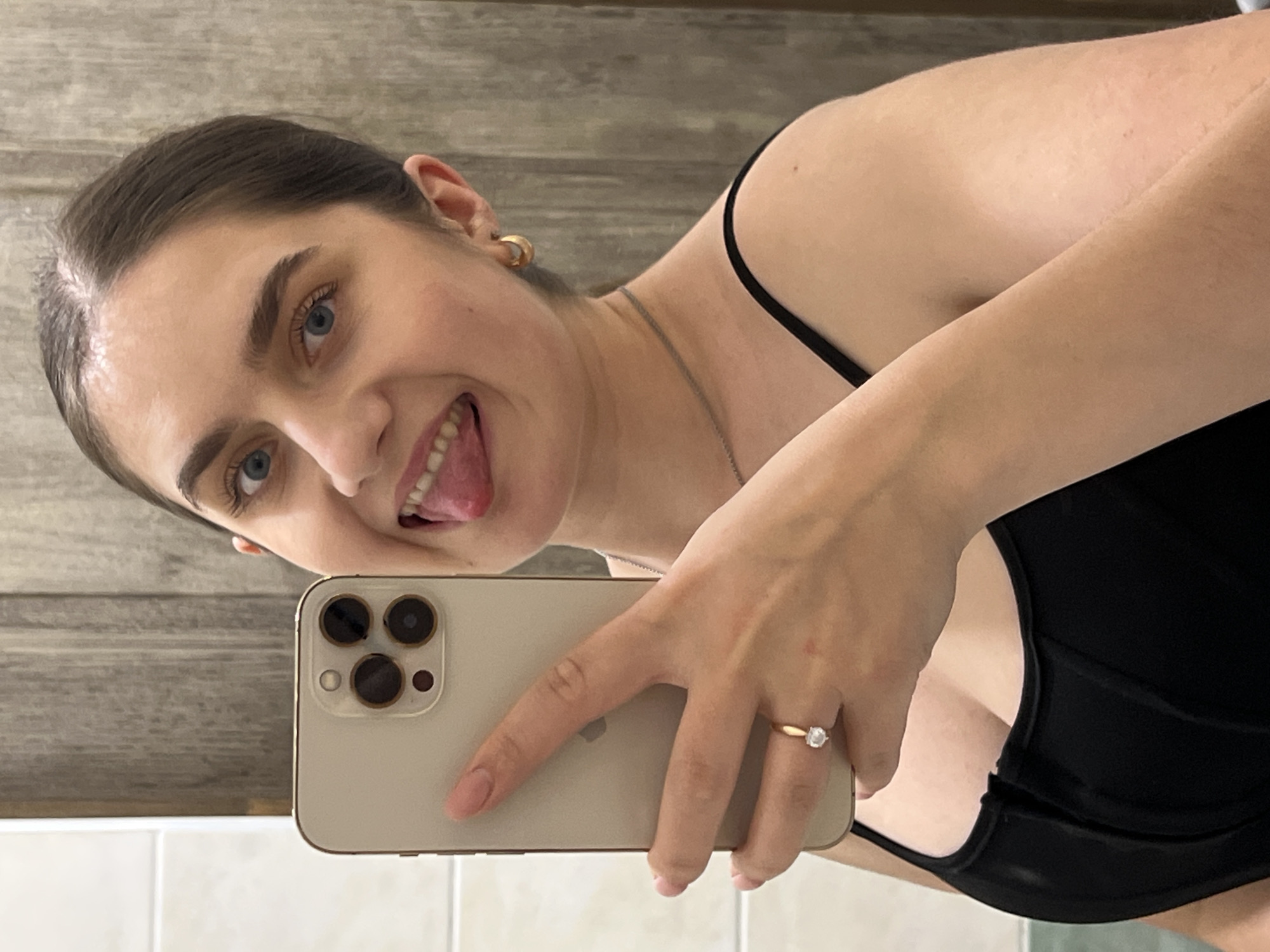 A young woman is taking a selfie indoors. The woman has fair skin, dark hair pulled back, and blue eyes. She is sticking her tongue out in a playful expression. She is wearing a black top with thin straps, a gold chain necklace, and gold hoop earrings. On her ring finger, she wears a gold ring with a prominent stone. She is holding a gold iPhone in her left hand. Behind her is a light-colored wall with a wood-like pattern. The bottom of the frame includes a section of white tiled wall. The location is not discernible from the image.