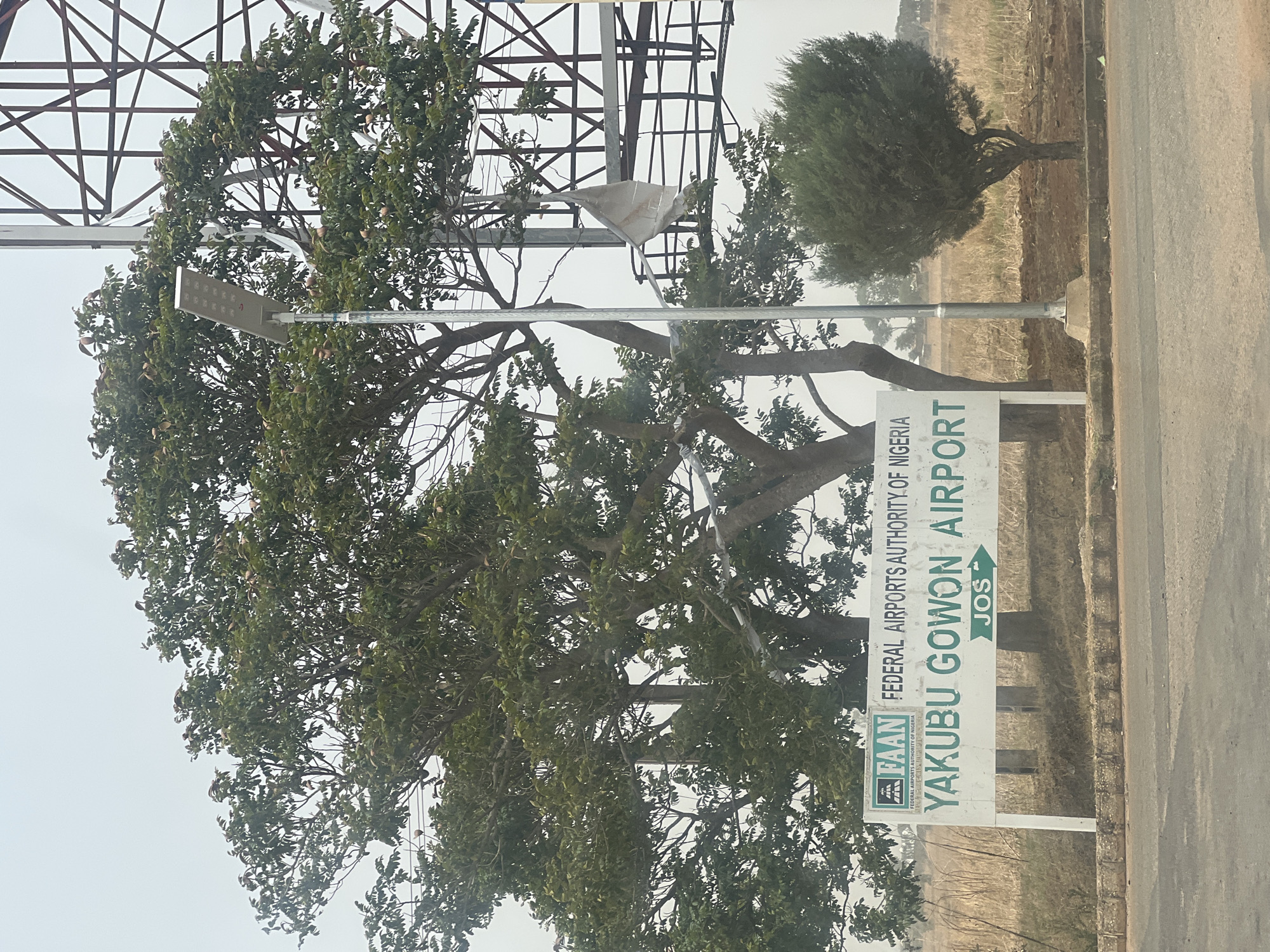 The image depicts a road sign indicating the location of Yakubu Gowon Airport in Jos, Nigeria. The sign, which is partially obscured by a large tree, also shows the logo of the Federal Airports Authority of Nigeria (FAAN). A metal lattice structure is visible behind the tree, and a small, rounded bush stands further down the road. The environment appears arid with dry grass. The image is somewhat distorted, suggesting it was taken at an angle.