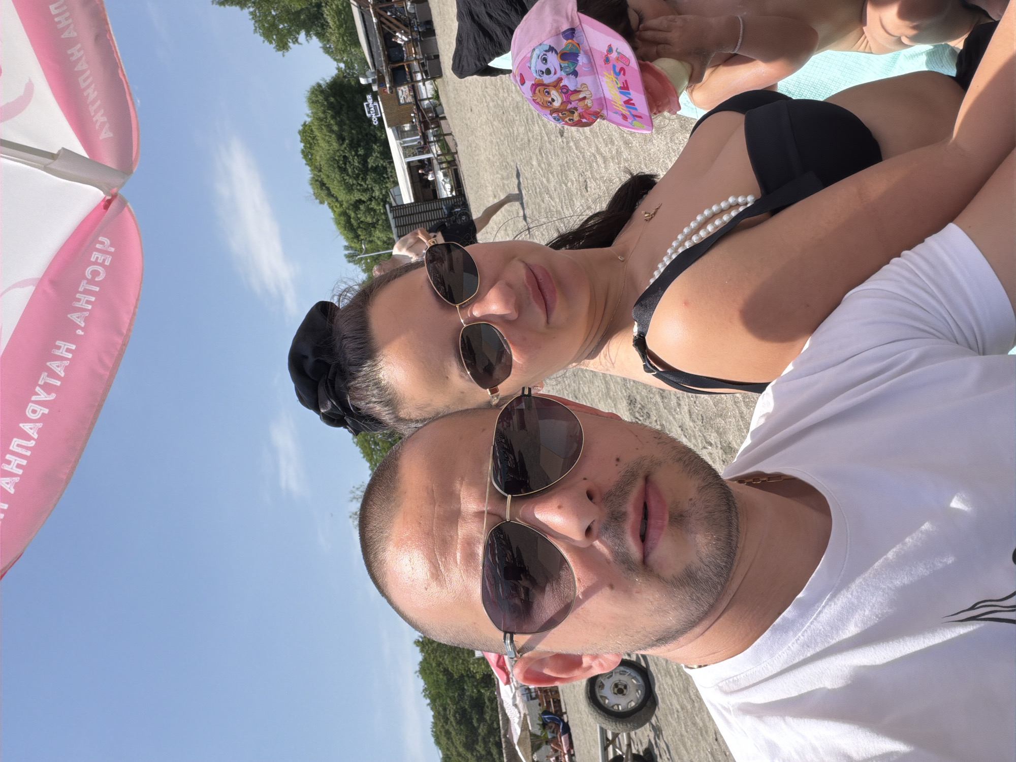 This image depicts three individuals relaxing on a sandy beach, identified as the Central Beach in Burgas, Bulgaria, based on the visible Cyrillic text "ПЛАЖЕН ХАТ" (Beach Hut) on a pink and white umbrella in the upper frame, indicative of a specific beach bar. The sky is clear blue with minimal clouds, suggesting bright, sunny weather.
In the foreground, a light-skinned adult male with a buzz cut and short beard wears aviator-style sunglasses and a white crew-neck t-shirt with a visible gold chain. He is holding the camera for a selfie, looking directly into the lens. To his right, a light-skinned adult female with dark hair tied in a high bun wears oval-shaped sunglasses, a black bikini top, a pearl-like necklace draped over her shoulder, and a delicate gold pendant necklace. She also looks towards the camera with a slight smile.
Partially visible to the right of the woman is a young child, wearing a pink baseball cap featuring "Paw Patrol" characters and the text "HAPPY TIMES". The child's right arm and shoulder are bare, and a thin bracelet is on its wrist. The child appears to be holding a light-colored object, possibly food, to its mouth while seated on a light blue towel or blanket.
In the mid-ground, a sandy beach extends with several distant figures, some under other beach umbrellas. Towards the right background, a structure labeled "CONTAINER" is visible, along with other wooden beach bar-style buildings. Green foliage and trees line the distant horizon, separating the beach from the adjacent area. On the far left, a portion of a cart or trailer with a wheel is present on the sand.