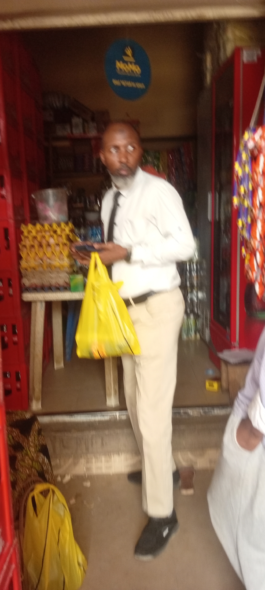 A man stands in the doorway of a small shop or kiosk in Fagge D2, Nigeria. He is bald with a dark beard, wearing a white long-sleeved shirt, a black tie, light-colored (khaki) pants, and black athletic shoes. He holds a yellow plastic shopping bag in his left hand and what appears to be a mobile phone in his right. His gaze is directed slightly to his right, away from the camera, with a neutral expression.
The shop interior is visible behind him, featuring red crates or shelving to his left. On a low table, several stacks of egg cartons are prominently displayed. Various other goods, including bottles and packaged items, are arranged on shelves further inside the shop. To his right, a red refrigerator is visible, and colorful strings of what appear to be packaged snacks or trinkets hang near the doorframe. The lower body of another person, wearing light-colored trousers, is partially visible on the far right. A discarded yellow plastic bag lies on the ground near the bottom left.
Above the man's head, a blue circular sign hangs, displaying the text "MoMo" in yellow lettering, with additional smaller, blurry text below it. The setting suggests a local retail business, likely active during the daytime.