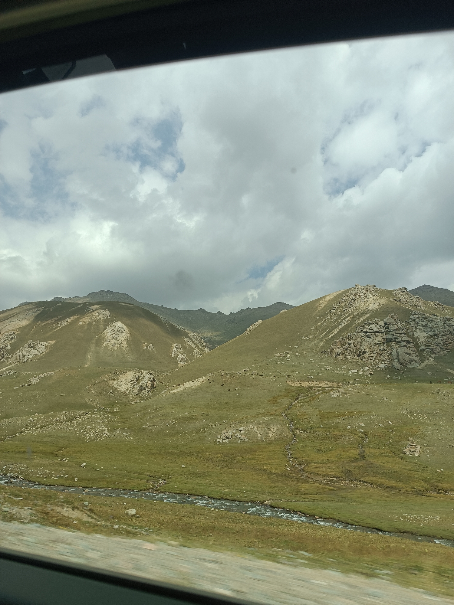 The image captures a wide, expansive view of a mountainous landscape in the Bala-Chychkan area of Kyrgyzstan. The terrain is characterized by rolling, grassy hillsides interspersed with large, rocky outcroppings. A narrow, winding river flows through the foreground, its water appearing clear and shallow. The sky above is partly cloudy, with dramatic, heavy clouds casting shadows on the mountains. There are a few small, dark figures visible on the grassy slopes, possibly livestock such as sheep or horses, indicating sparse vegetation and grazing activity. The overall impression is one of a natural, relatively undeveloped wilderness with rugged beauty.
