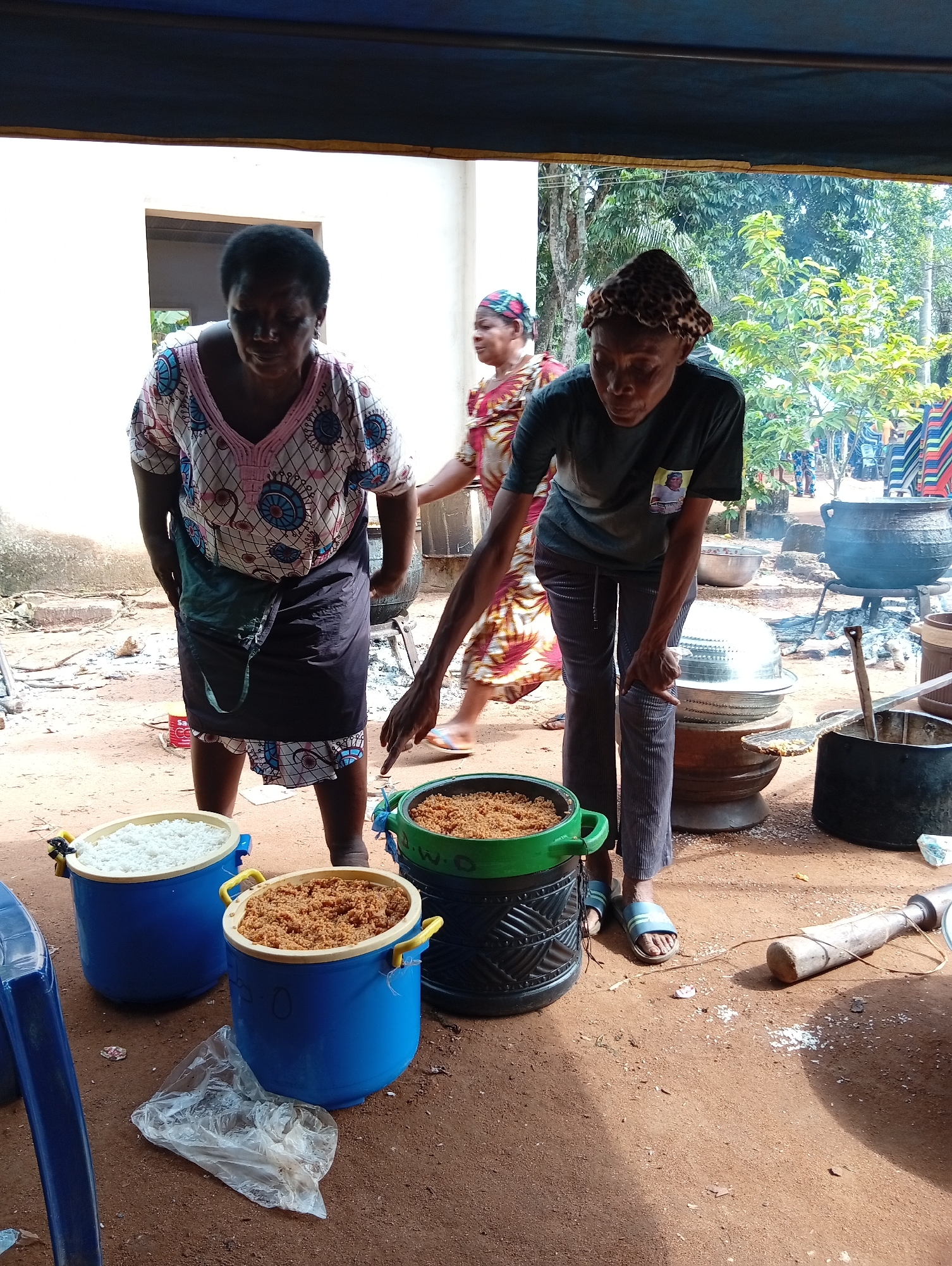 The image depicts an outdoor scene of food preparation or serving, likely at a communal gathering or event. In the foreground, two women are actively engaged. The woman on the left, wearing a white and pink patterned top with blue circular designs and a black skirt, bends forward looking downwards. To her right, another woman, sporting a dark t-shirt with a small pinned photo on the pocket, dark trousers, and a patterned headscarf, is also bent over, seemingly attending to a large green and black patterned bucket. She wears light blue slide sandals.
Between these women are three large containers of food. Two blue plastic buckets with yellow handles hold white rice and a reddish-brown granular substance (possibly garri or a similar grain), respectively. The green and black patterned bucket also contains the reddish-brown substance. Faint text "W-O-" is visible on the green bucket, and "C-O" on one of the blue ones.
In the mid-ground, another woman in a patterned dress walks past. The ground is reddish-brown dirt. In the background, a light-colored building and lush green trees and foliage indicate a bright, sunny daytime. Large black cooking pots are visible, one on a metal stand over what appears to be an open fire, suggesting traditional cooking methods. A large wooden pestle lies on the ground. Stacks of colorful plastic chairs in the distant background further hint at a public or festive event. A clear plastic bag lies on the ground near the food containers. The scene conveys a sense of busy activity centered around preparing and distributing food for a gathering.
