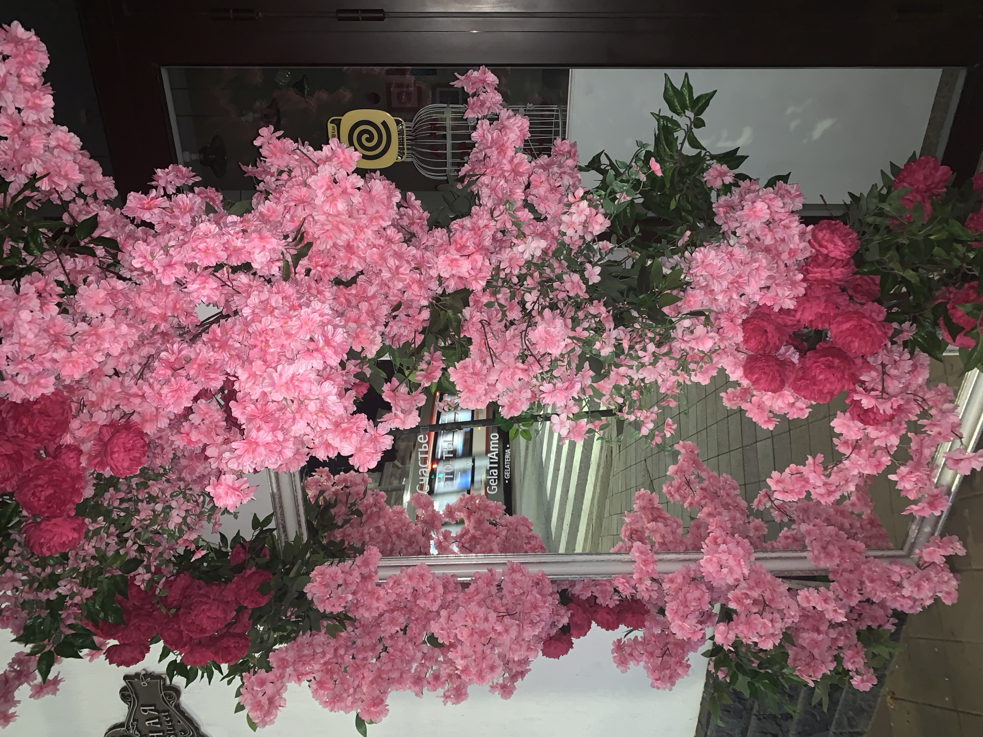 A medium shot captures a vibrant display of artificial pink and fuchsia flowers cascading around a mirrored surface and a dark brown doorway. The flowers are densely packed, creating an archway effect. To the left of the arch, a white wall with a dark grey stone base is visible. A decorative metal sign with ornate swirls and Cyrillic text reading "Улица" ("Street") and "Street" in English is attached to the white wall.
The mirrored surface reflects a crosswalk, a streetlamp, and the text "Счастье, то что ты" ("Happiness, that you are") and "GELATIAMO GELATERIA" on a storefront. A yellow sign with a black spiral design resembling a stylized jar or a cage is affixed to the glass door of the establishment. The scene appears to be an outdoor storefront decorated for an event or ambiance. The lighting suggests it is evening or nighttime.