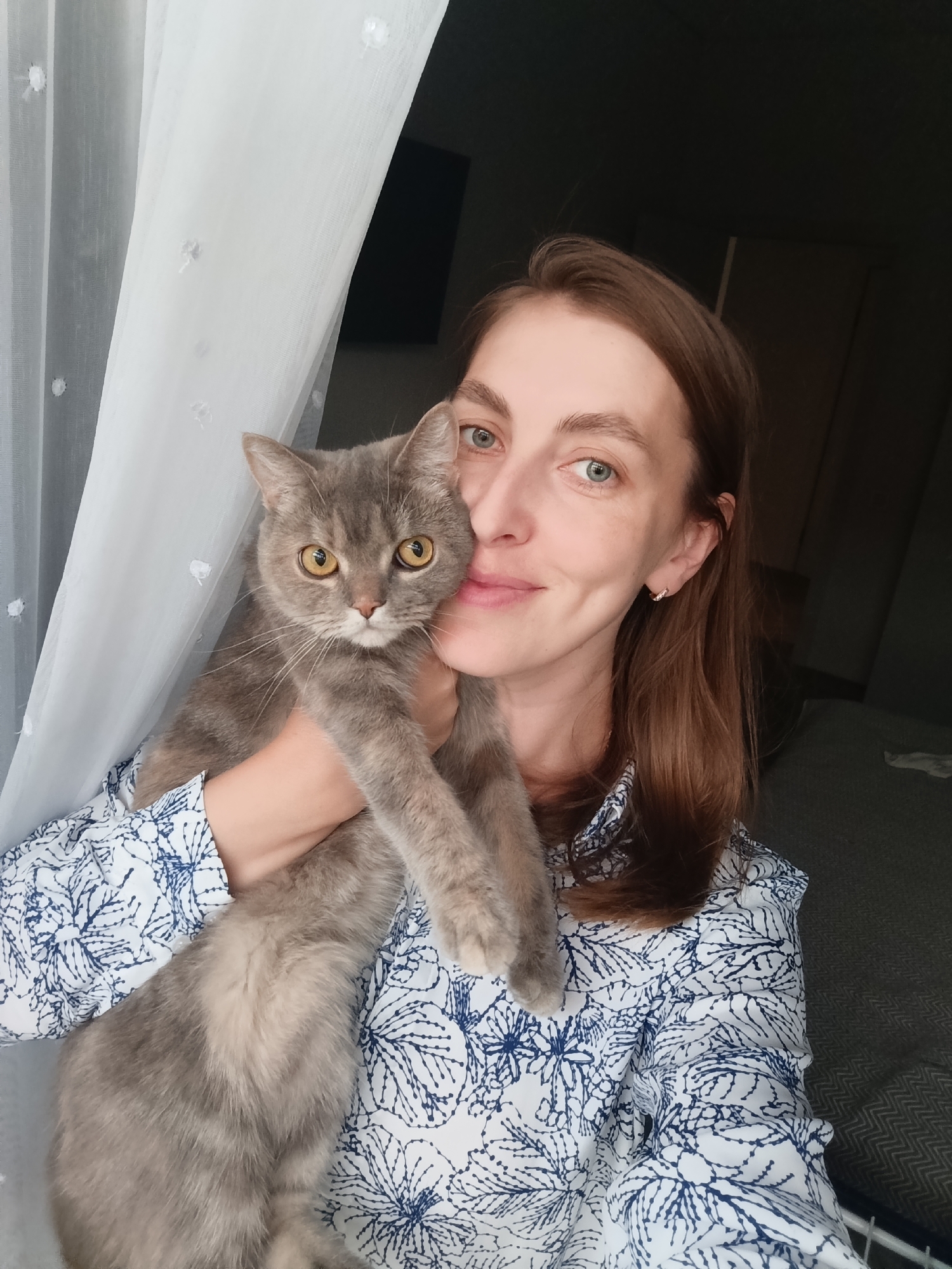 In a home setting, likely an apartment in Chaikovsky, Russia, a woman with shoulder-length brown hair and blue eyes is captured in a selfie. She is wearing a white blouse with a blue floral pattern. The woman is holding a grey cat with yellow eyes close to her face. The background shows a sheer white curtain on the left and a dark wall with some furniture visible on the right. The lighting suggests it is daytime.
