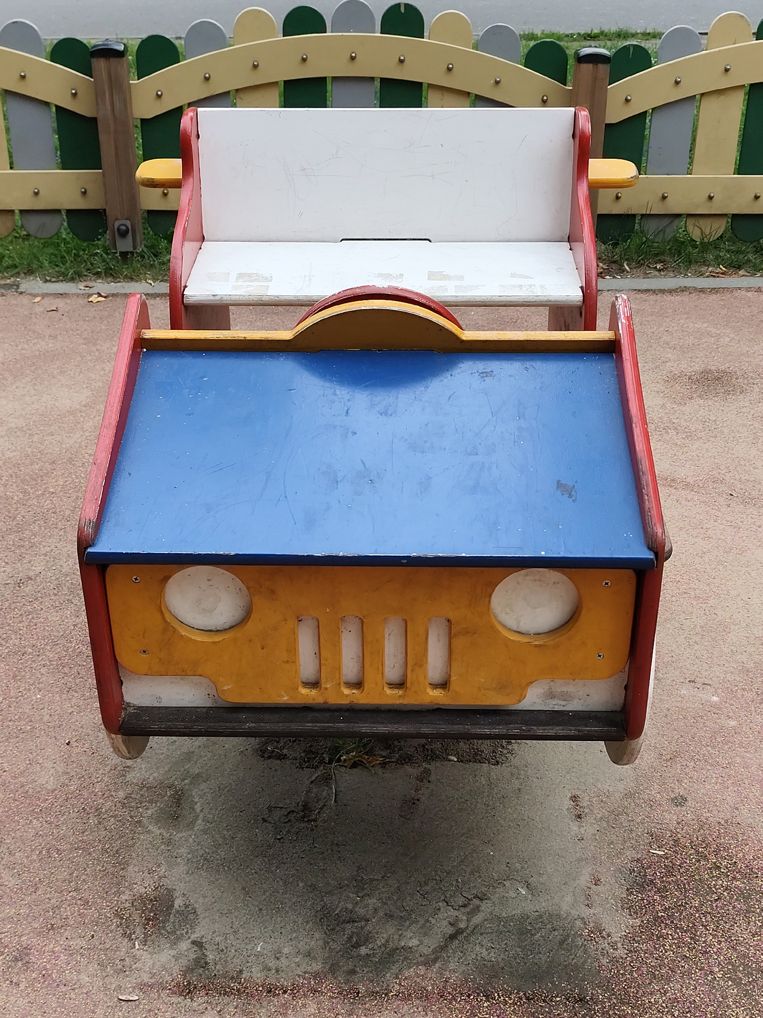 The image shows a wooden playground ride shaped like a car. The car is red, yellow, and blue and has white seats. It is located on a playground with a fence in the background. The ground is covered in red gravel and some dirt. The image does not provide any information about its location in Moscow, Russia.