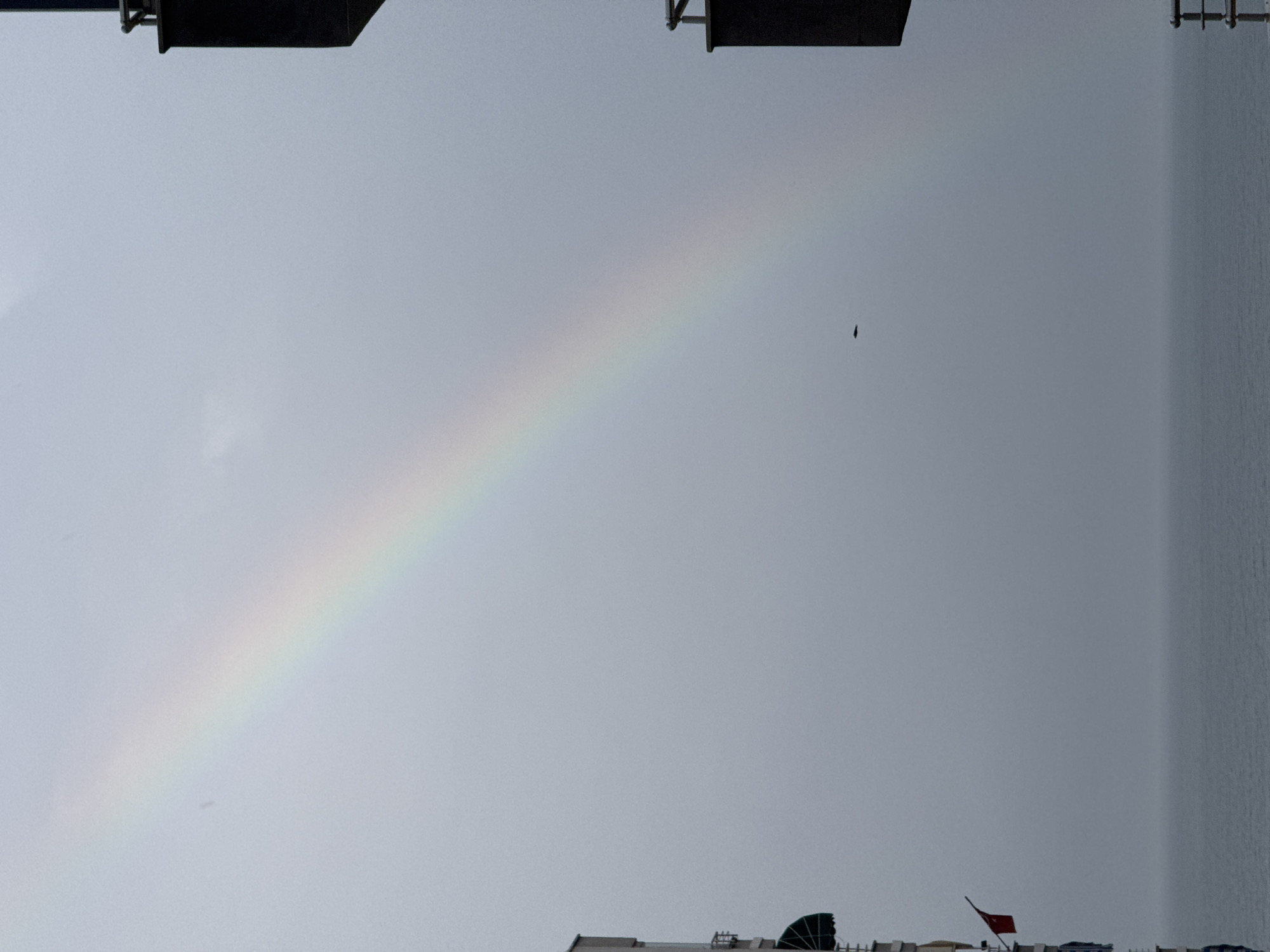 The image depicts a rainbow in a cloudy sky over what appears to be a section of Erdemli, Turkey. In the foreground, the tops of buildings with various structures and a small red flag are visible. Above, a faint rainbow stretches across the sky, slightly left of center, and a bird is flying in the sky, to the right of the rainbow. The top of the frame shows partial images of dark-colored structures or objects, possibly parts of buildings or street fixtures.