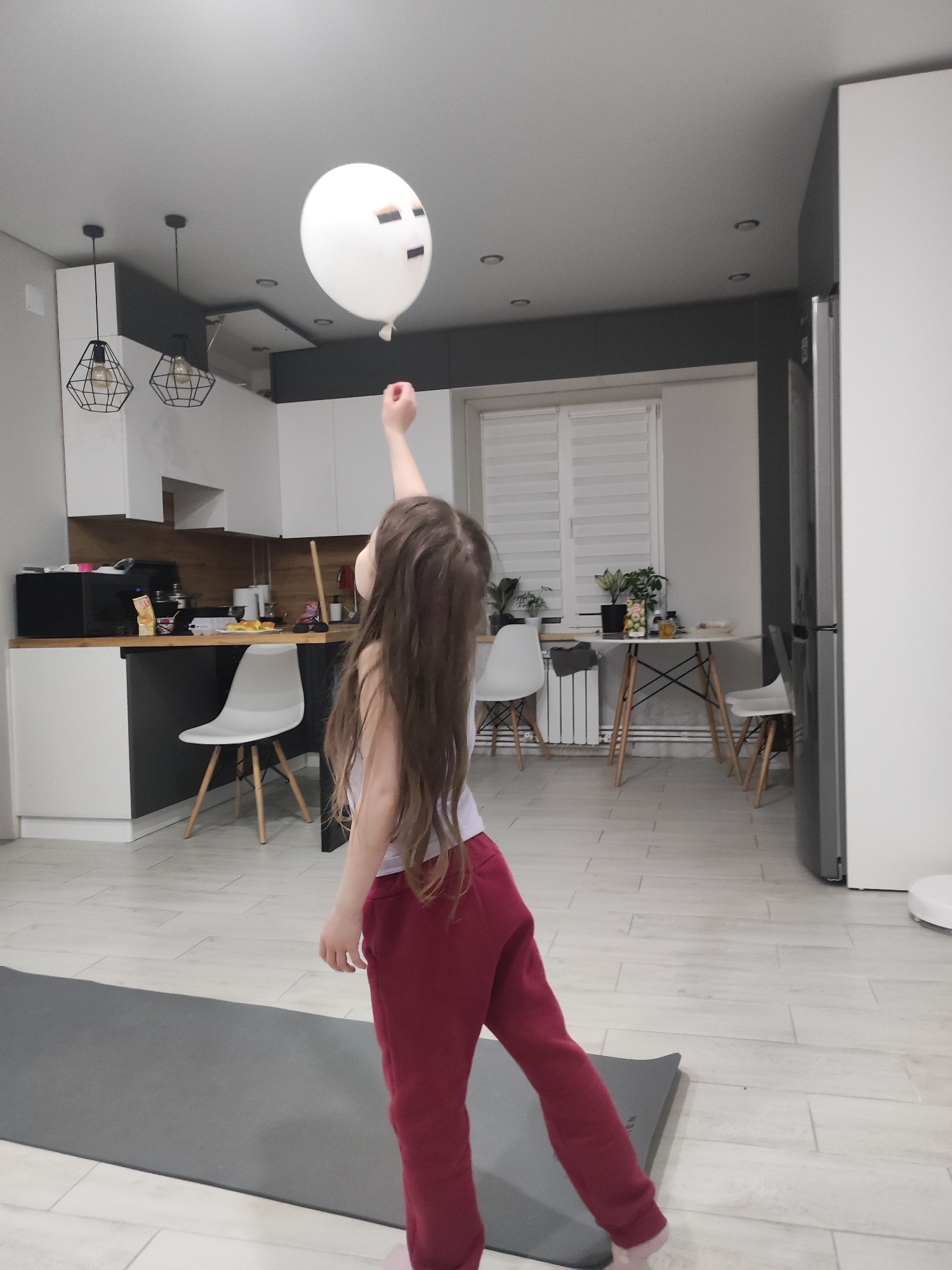 A young girl with long brown hair, wearing a white shirt and red pants, reaches for a white balloon with a drawn-on face. The scene is an interior, likely a modern apartment, featuring a kitchen area with white cabinets, a black countertop, and a dining area with a white table and chairs. A gray yoga mat lies on the light-colored tile floor. The location is impossible to determine definitively from the image alone, and there is no definitive connection to the city of Votkinsk, Russia.