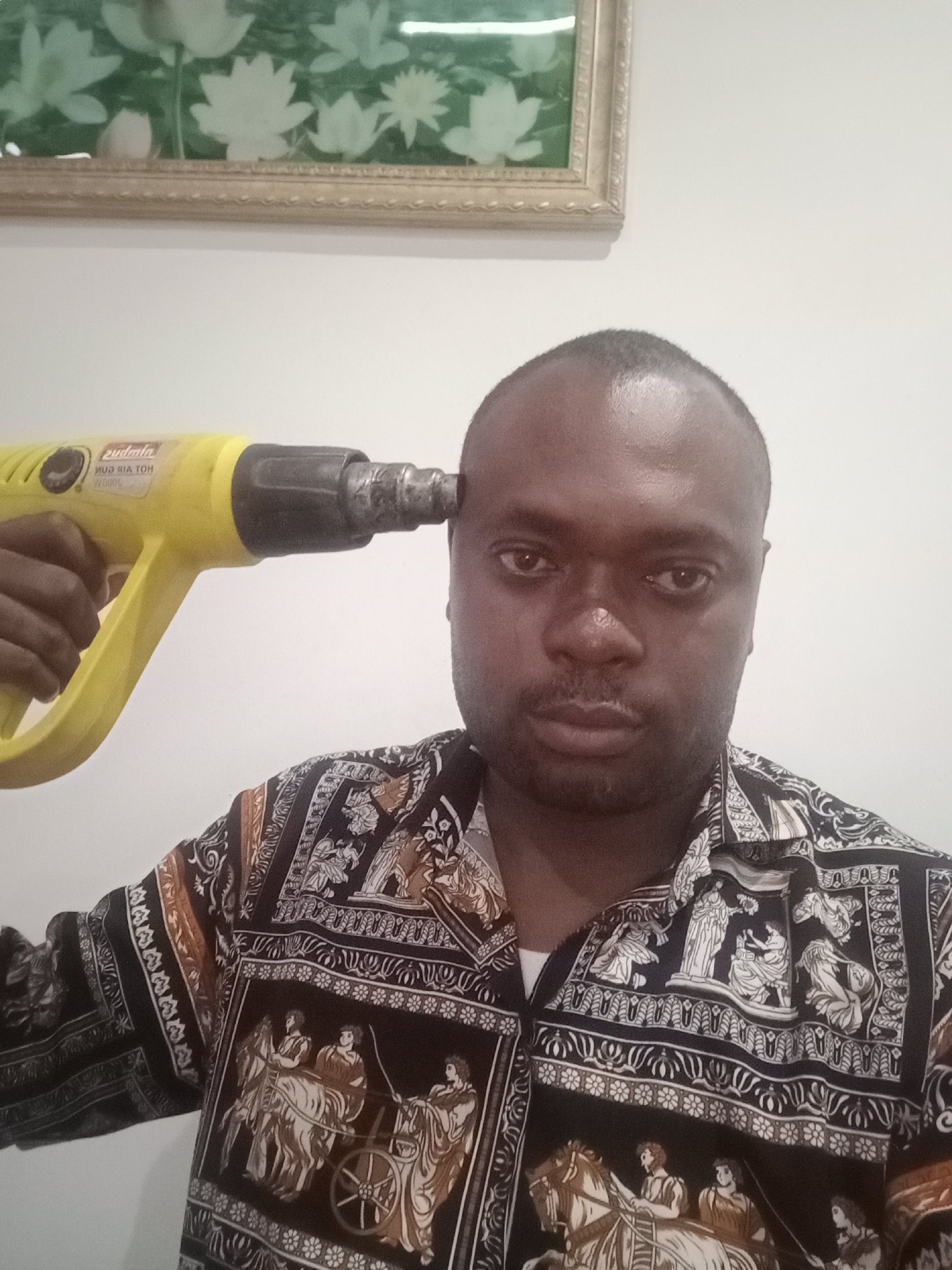 A dark-skinned man with a shaved head and a faint mustache/beard is the central subject of the image, captured in what appears to be an indoor setting in Gwarinpa, Nigeria. He wears a patterned short-sleeved shirt featuring classical-style imagery in black, white, and brown, depicting figures in chariots, on horseback, and engaged in various scenes, bordered by intricate patterns.
The man holds a bright yellow and black heat gun to his right temple, mimicking the gesture of pointing a firearm. He looks directly into the camera with a serious and unsmiling expression. The background consists of a plain white wall, with a framed picture visible in the upper left corner. This picture contains a green background with white, lotus-like floral designs.
The lighting is bright and even, suggesting either strong artificial light or natural daylight within the indoor space. Visible text on the heat gun, though partially reversed, includes "HOT AIR GUN" and "2000W," along with what might be a brand name like "Sudata." The overall mood conveyed by the man's expression and the pose with the heat gun is one of directness or possibly a symbolic, theatrical gesture rather than genuine distress.