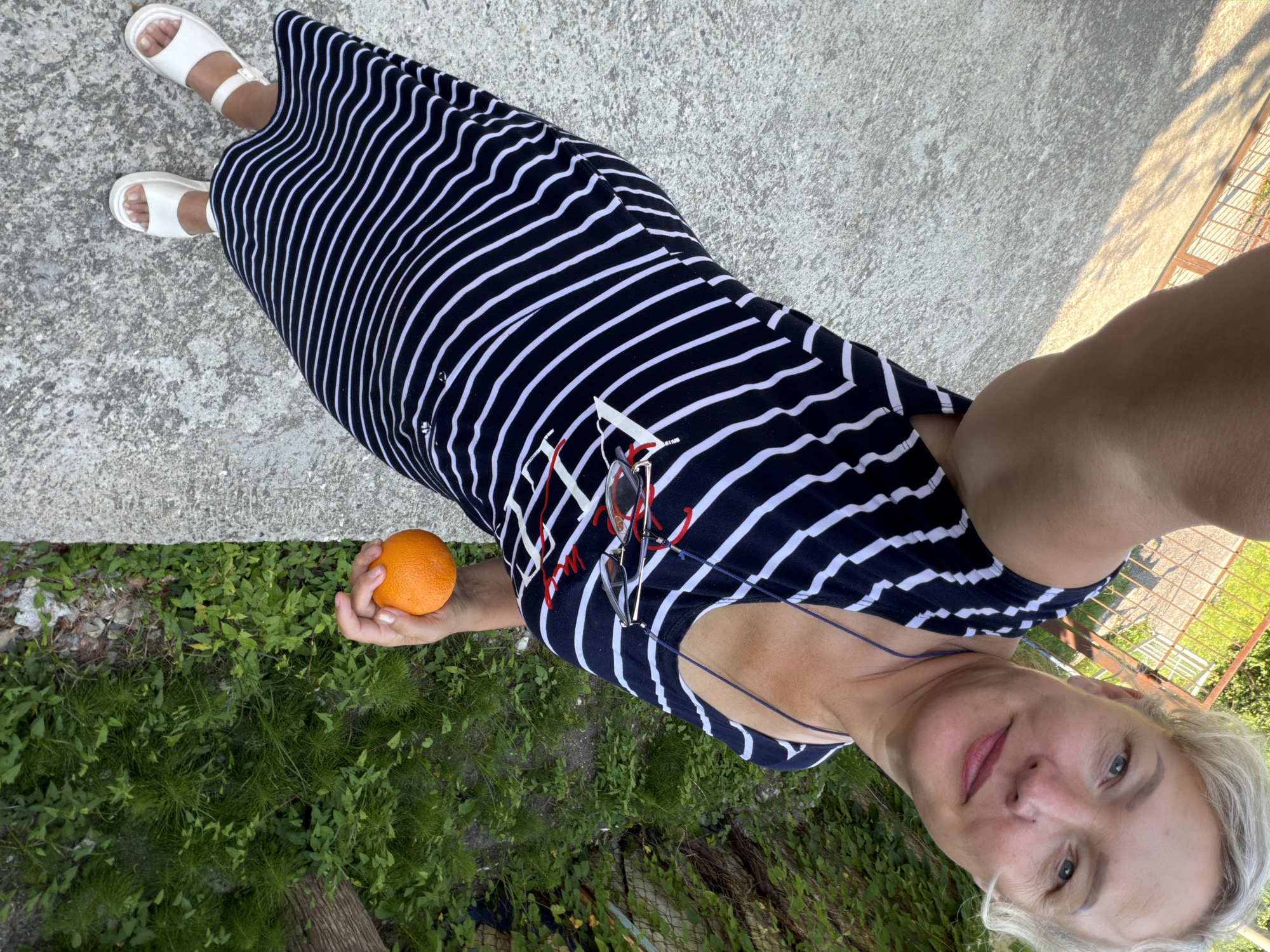 A woman with blonde hair is standing on a concrete surface in an outdoor setting. She is wearing a black and white striped dress and white sandals. She is holding an orange in her right hand. In the background, there is lush green vegetation. The image appears to have been taken in Shepsi, Russia, based on the context provided.