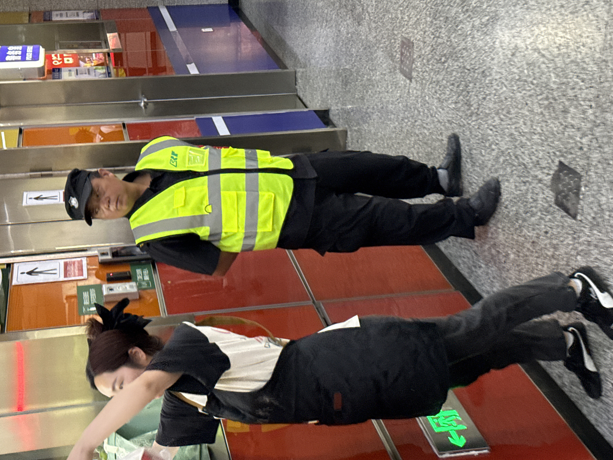 In an indoor setting, possibly a subway station in Yuzhong District, China, two people are visible. The first person is wearing a black uniform and a bright yellow reflective safety vest, along with a dark cap. They are lying prone on the floor near a wall with red and orange panels and informational signs. The second person, possibly a food vendor, is lying in a similar prone position, dressed in a white shirt with black sleeves and dark pants. A green exit sign is adjacent to them on the wall. The floor is speckled grey.