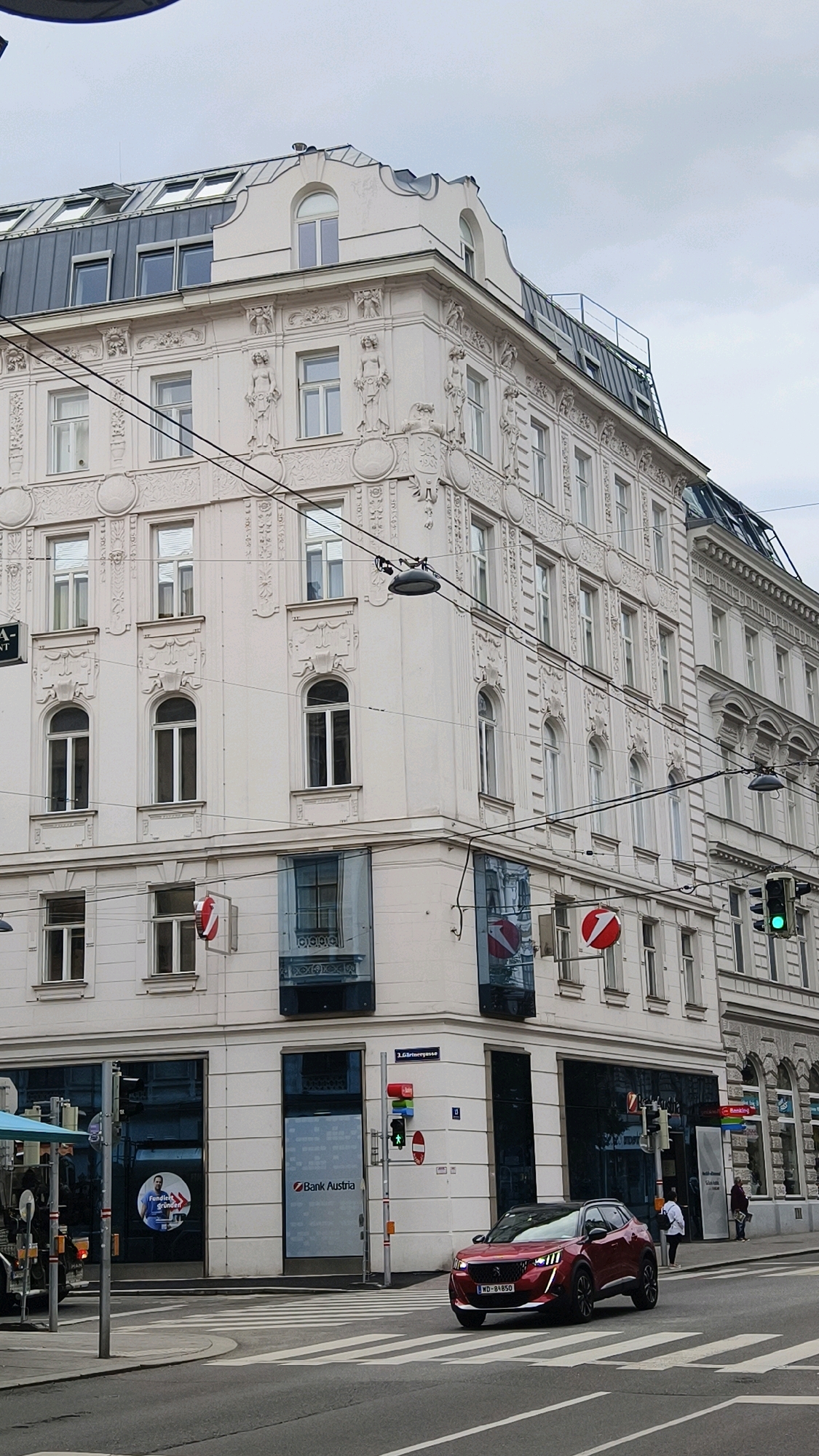 The image captures a street view of a corner building in Vienna, Austria, under an overcast sky. The building is an ornate, multi-story structure with light-colored facade, featuring numerous windows and decorative architectural elements. The lower levels house a Bank Austria branch, identifiable by its signage and reflective glass windows.
In the foreground, a modern red car is driving across a pedestrian crossing. The car has a license plate that reads "WD-84850". Traffic lights are visible, indicating green for the car. A traffic sign with a white circle and a red diagonal line is also present.
The street is paved and includes a crosswalk. On the left side of the image, part of a blue awning and a street pole are visible, suggesting a bus stop or similar urban amenity. A few pedestrians are present on the sidewalks in the background, near the entrance of the bank and further down the street. Overhead wires for what appear to be trams or streetcars crisscross the scene. The overall impression is of an active urban environment in Vienna.