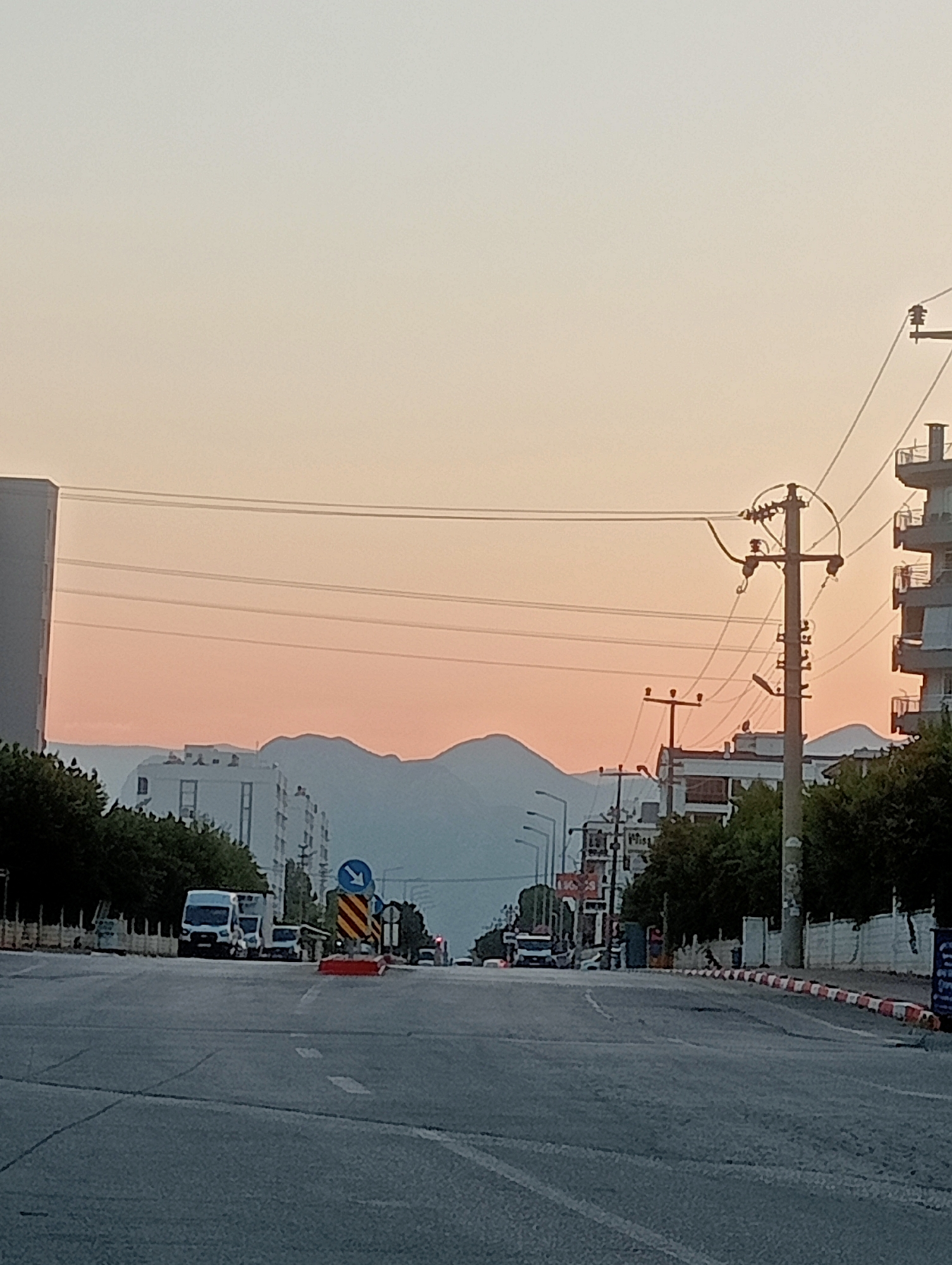 The image shows a street scene in a city at dusk. The sky is a gradient of pale orange and pink. In the foreground, a road is visible with lane markings. To the right, a red and white striped traffic cone is placed on the side of the road. Beyond the cone, there are buildings and trees. In the distance, mountains are silhouetted against the sky. Electrical poles and wires are prominent in the mid-ground and extend across the scene. Several vehicles, including a white van, are visible on the road. The overall impression is of a quiet street in a city during the evening hours.