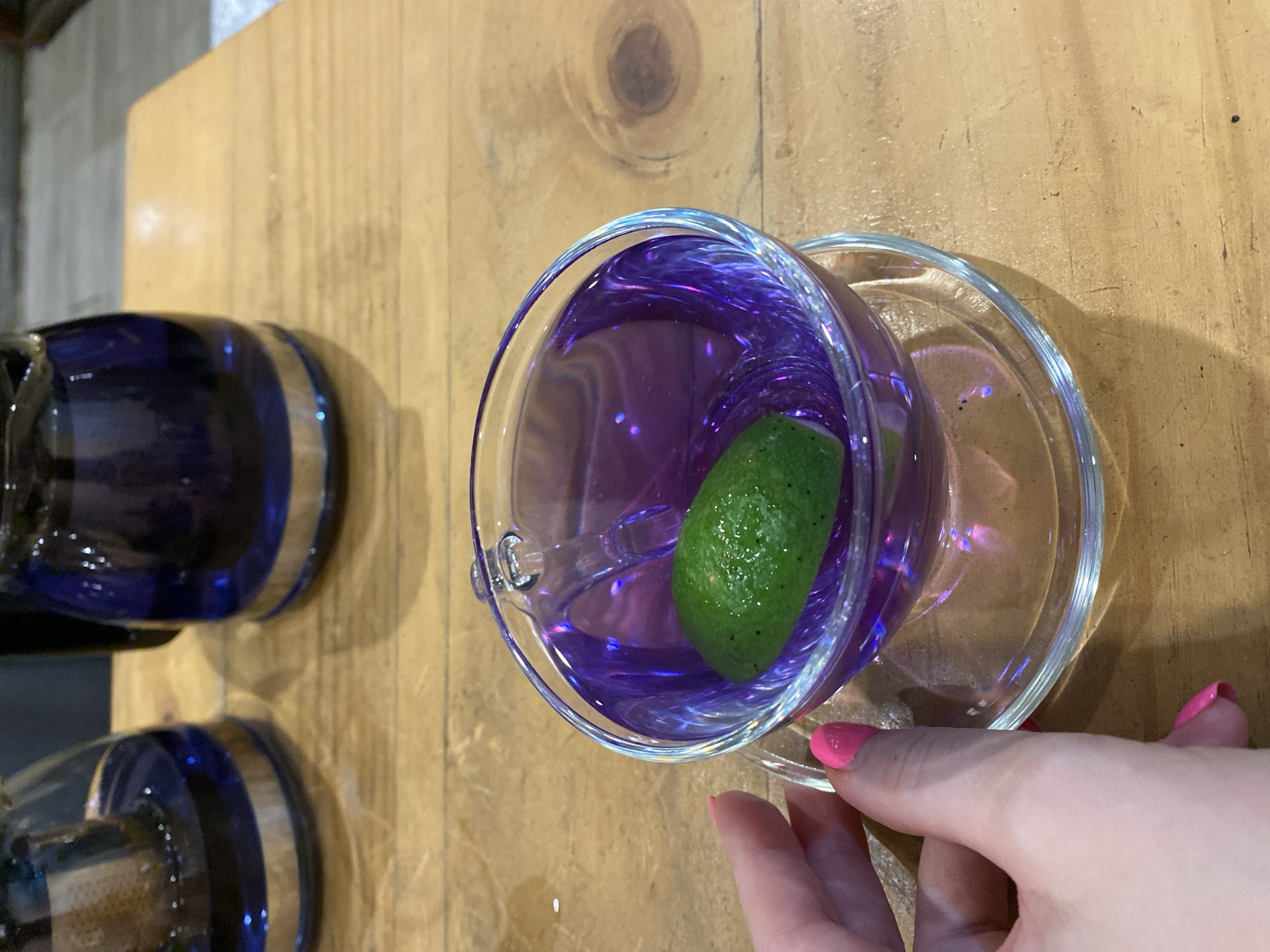 A hand with bright pink fingernails is reaching for a clear glass teacup filled with a vibrant purple liquid and a lime wedge. The teacup sits on a matching glass saucer, both placed on a light brown wooden table. In the background, out of focus, are two dark blue glass teapots, one with a silver infuser visible. The scene suggests a leisurely moment, possibly at a cafe or a home, in Da Nang, Vietnam.