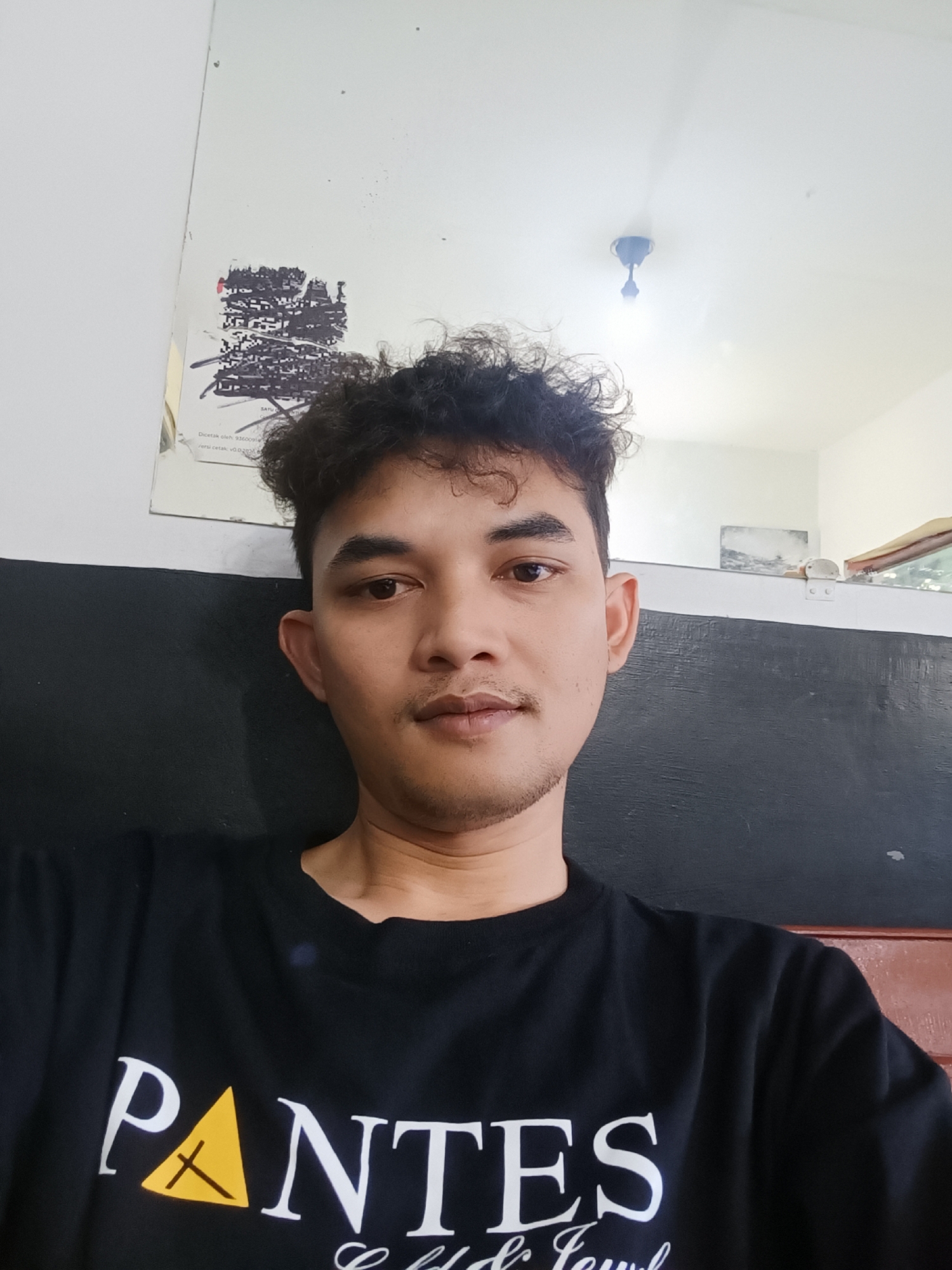 A medium-shot selfie captures a man with short, dark curly hair, light brown skin, and a trimmed mustache. He wears a black t-shirt with the word "PANTES" printed on it in white letters with a yellow triangle.
The background consists of a wall divided horizontally into a white upper section and a black lower section. Above the man's head is a mirror with a faded black and white graphic attached to it. A portion of the ceiling with a light fixture is visible in the mirror's reflection.
The scene is indoors. Given the prompt, the location is presumed to be somewhere in the city of Cijoho, Indonesia. No specific details in the image confirm this location.