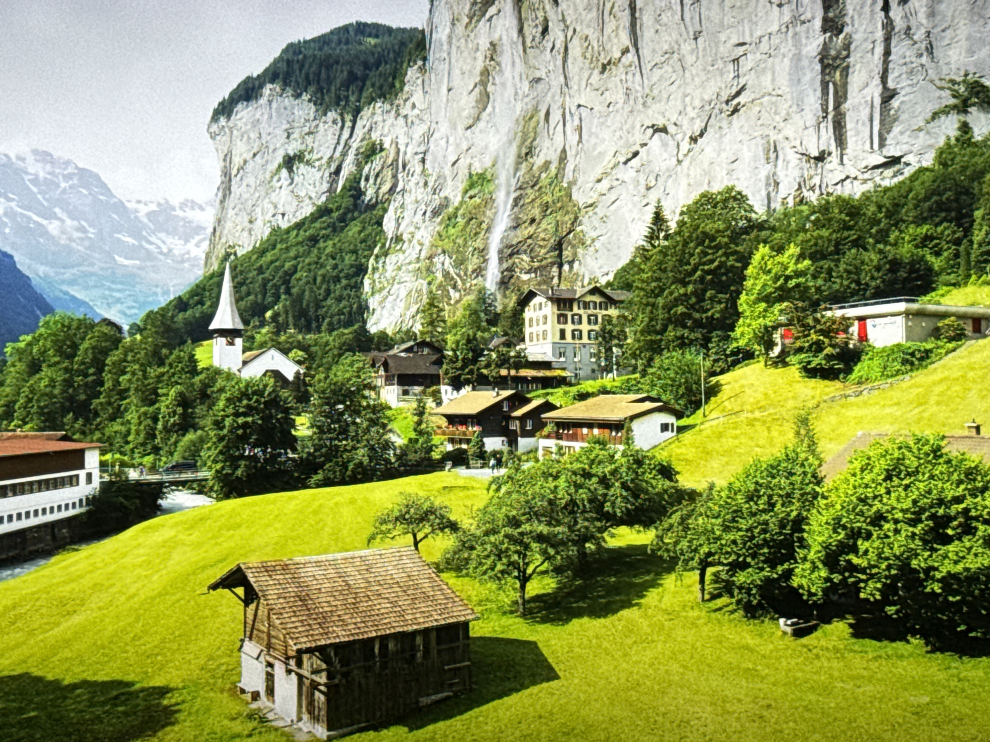 The image depicts a picturesque alpine village nestled at the base of a towering, sheer cliff face. A prominent waterfall cascades down the cliff. The village features traditional Swiss architecture with chalets and a church with a steeple. Lush green meadows slope upwards towards the mountains, dotted with trees. In the foreground, a rustic wooden barn sits on the grassy hillside. The scene is bathed in natural daylight. The image is a landscape photograph. The location is not Burgas, Bulgaria.