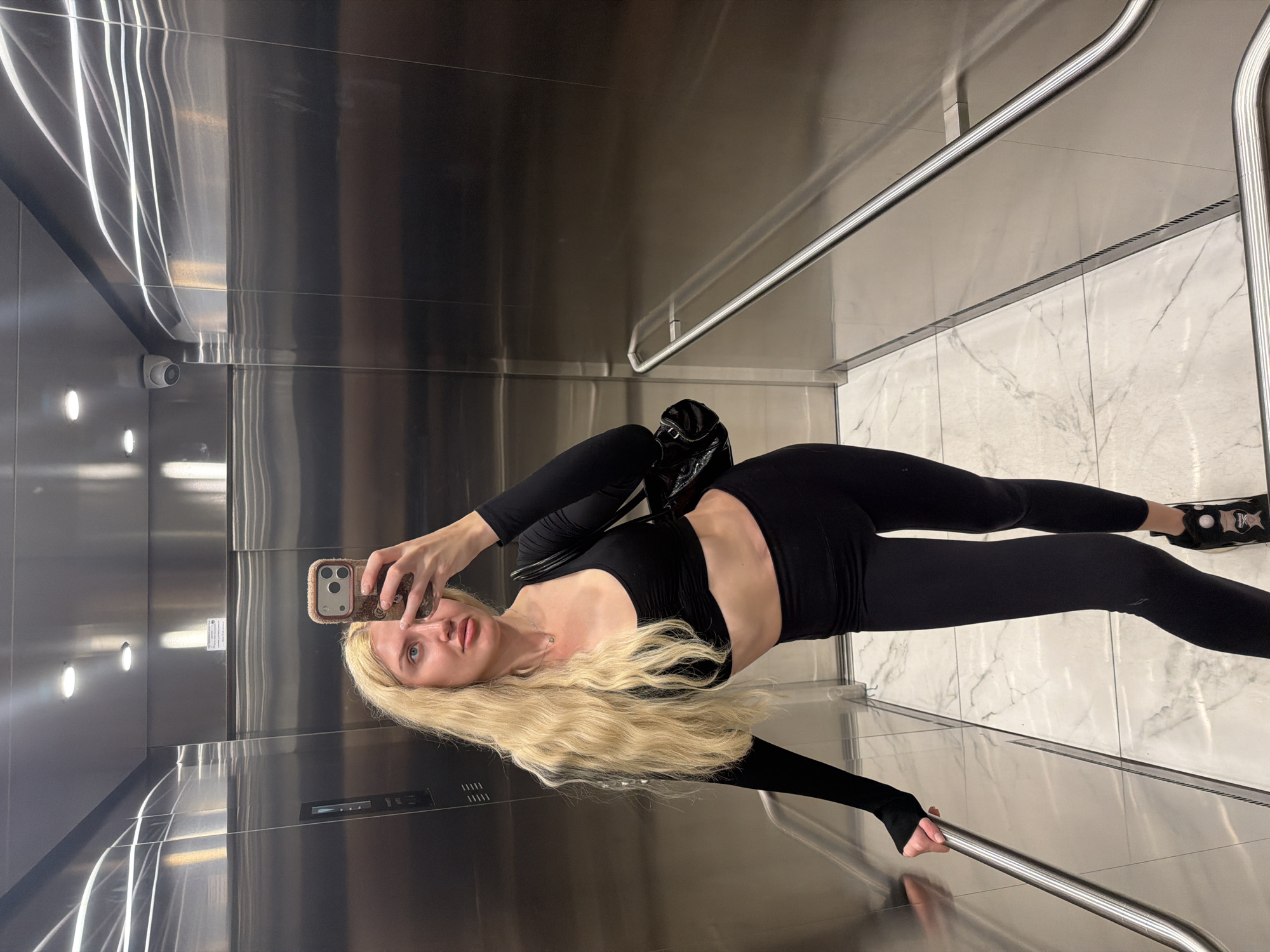 A blonde woman, wearing a black crop top and black leggings with black sneakers, poses for a selfie in an elevator. She is holding a cell phone in her right hand while leaning against the left wall of the elevator. She is also holding onto a horizontal grab bar with her left hand. The elevator's walls are metallic with vertical lighting, and the floor appears to be marble. A small black handbag is visible on her left side. A security camera is visible in the upper left corner. The location is likely in Belgrade, Serbia.