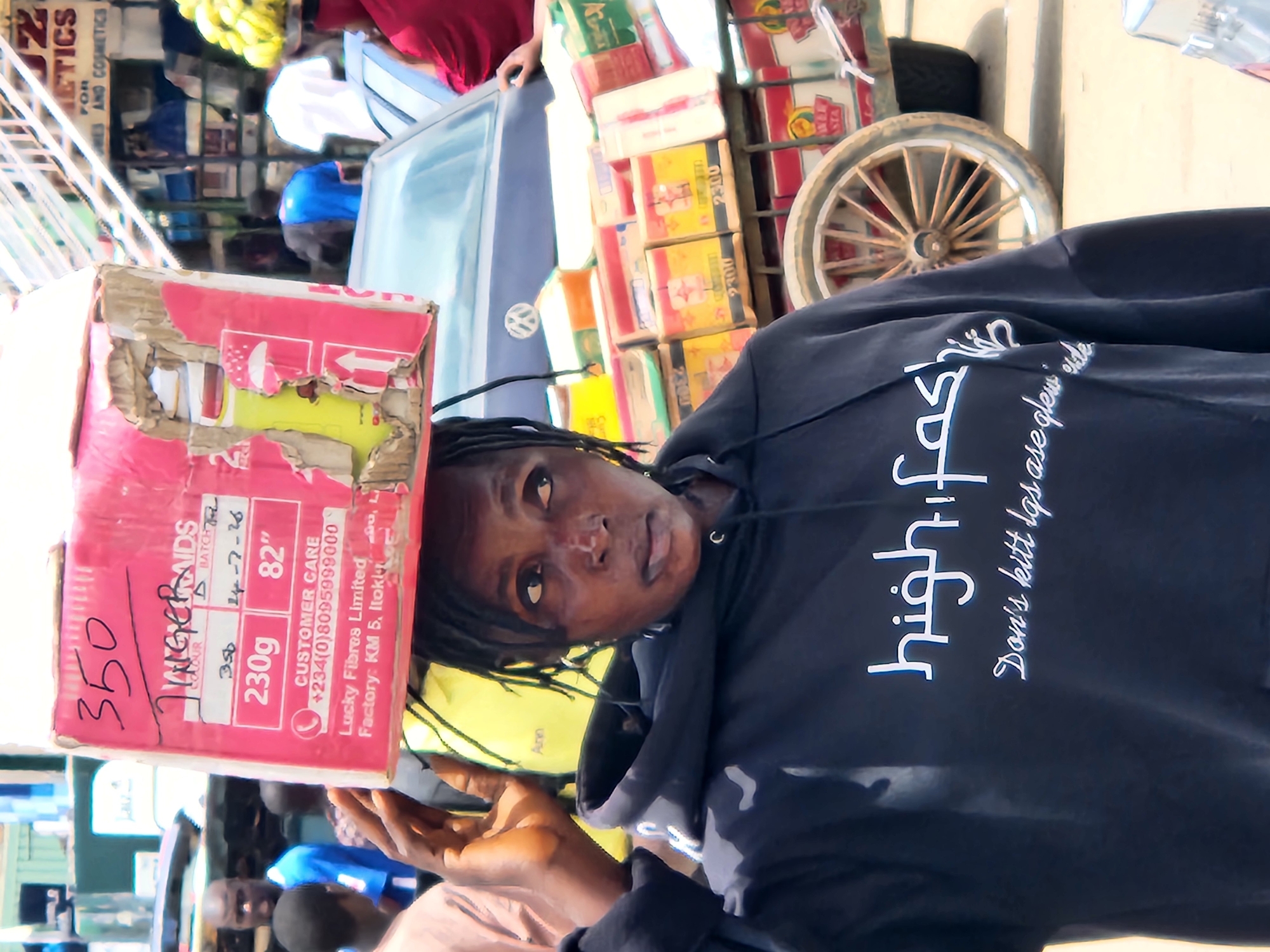 A woman balances a large pink cardboard box filled with hair products on her head, walking down a street in Jos, Nigeria. The box has handwritten labels with "350" and "JINGEBRAIDS" visible. The woman wears a dark hooded sweatshirt with white text that reads "high fashion" and "Don't kill the glow inside." Behind her, a cart is loaded with more boxes of hair products, a car, and other pedestrians. In the background, buildings with signs are visible, including one for "COSMETICS." The scene appears to be a busy marketplace.