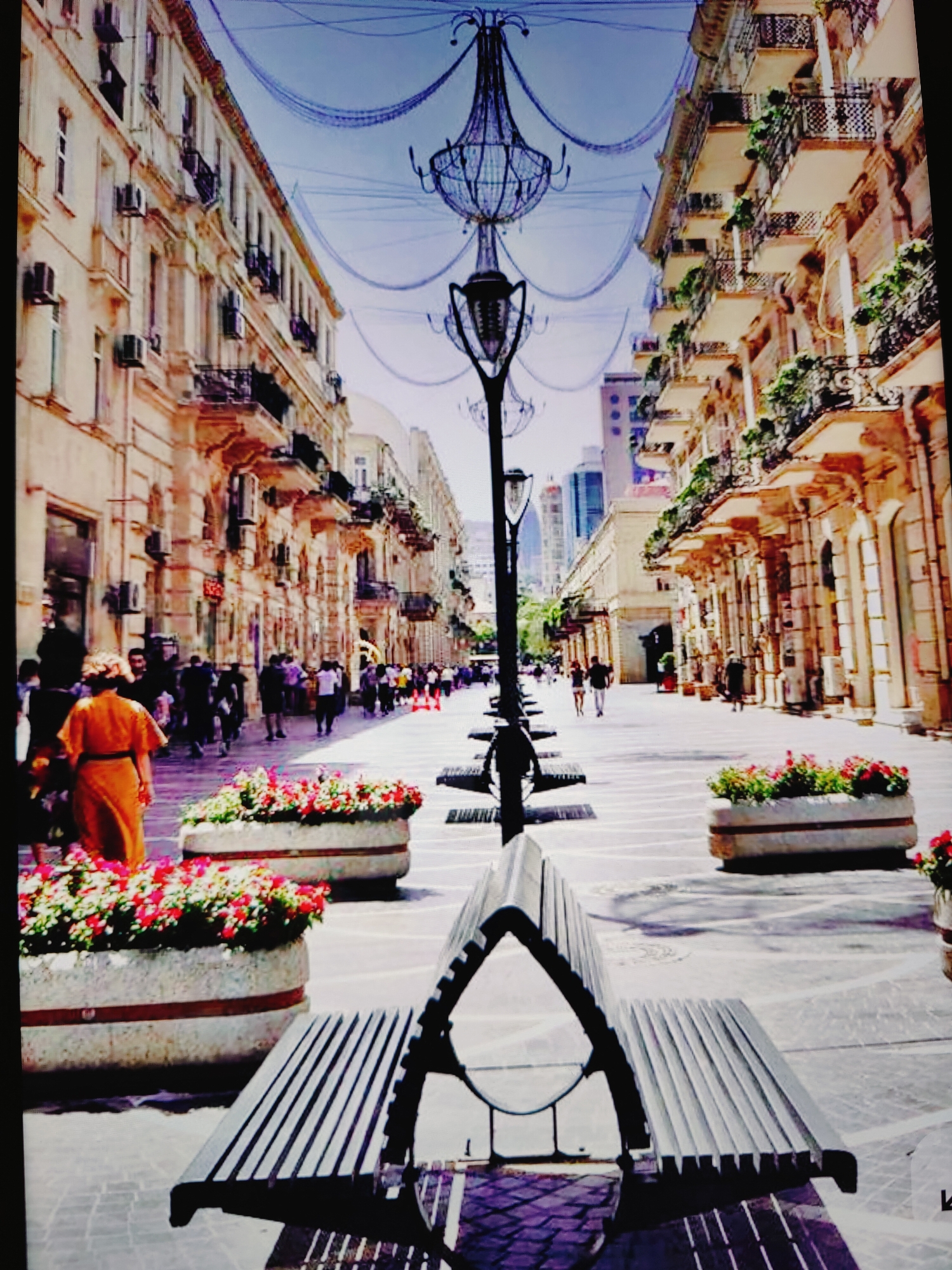 The image depicts a pedestrian street scene in Baku, Azerbaijan, likely Nizami Street, known for its ornate architecture and lively atmosphere. In the foreground, a modern metal bench with a curved backrest is positioned on a patterned stone walkway. Behind the bench, two large, rectangular concrete planters overflow with vibrant red and pink flowers, adding a splash of color to the scene.
Further down the street, people are strolling, some in groups and others individually. The buildings lining the street are multi-story structures with classical architectural elements, featuring balconies with decorative iron railings and ornate facades. Air conditioning units are visible on some of the buildings. Overhead, a decorative light fixture, resembling a large chandelier, is suspended by cables, along with more elaborate string lights that create an artistic canopy across the street. The sky above is a clear blue. The overall impression is one of a well-maintained, upscale urban environment, popular with both locals and tourists.