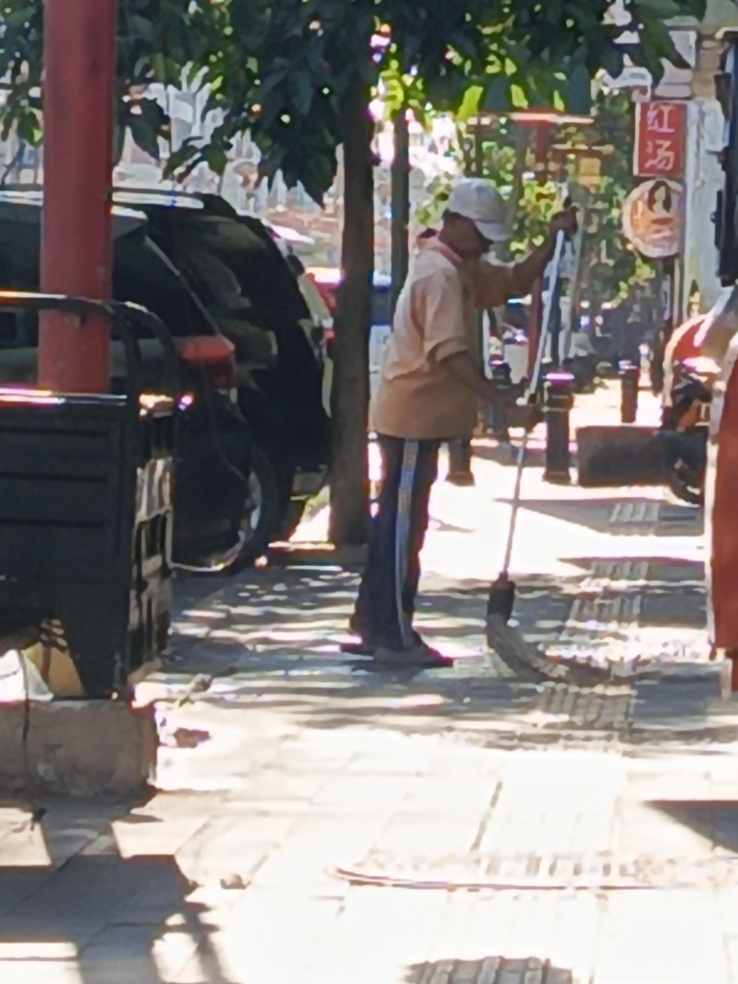 A person, wearing a light-colored short-sleeved shirt and dark pants with white stripes, is sweeping the sidewalk with a broom. The person is also wearing a white cap. The scene appears to be on a street in Surabaya, Indonesia. There are trees and buildings in the background. A black vehicle is parked on the left side of the image, and a red structure is visible to the left of the person. The ground is paved with tiles, and shadows from the trees are cast across the sidewalk.