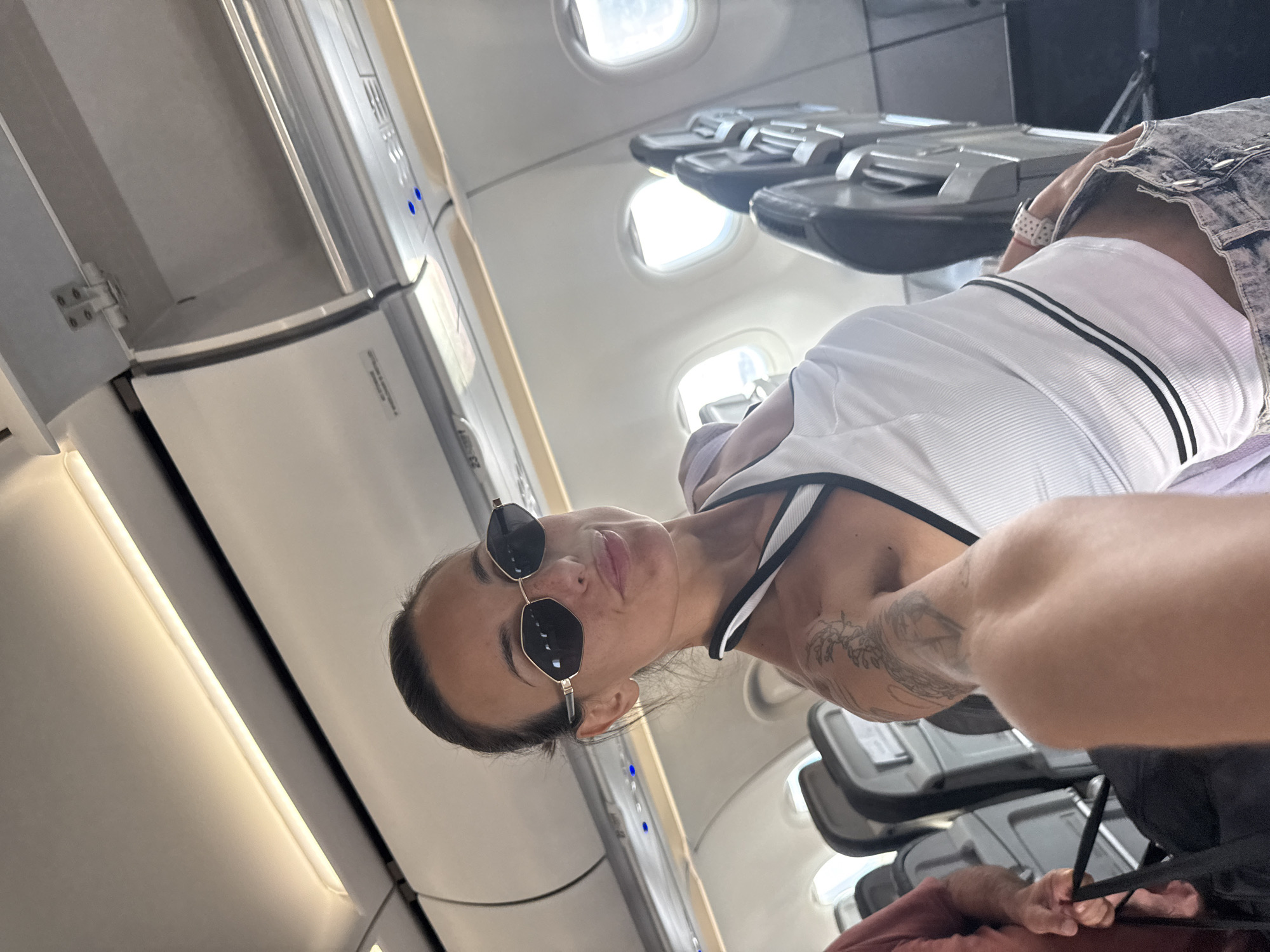 A woman in sunglasses and a white ribbed tank top is taking a selfie on an airplane. She is in the foreground, with rows of empty airplane seats visible behind her. To her left, a man in a red shirt is holding a black bag. The interior of the airplane is visible, including overhead compartments and windows. The lighting suggests it is daytime. There is no indication of the city of Sofia, Bulgaria, in the image.