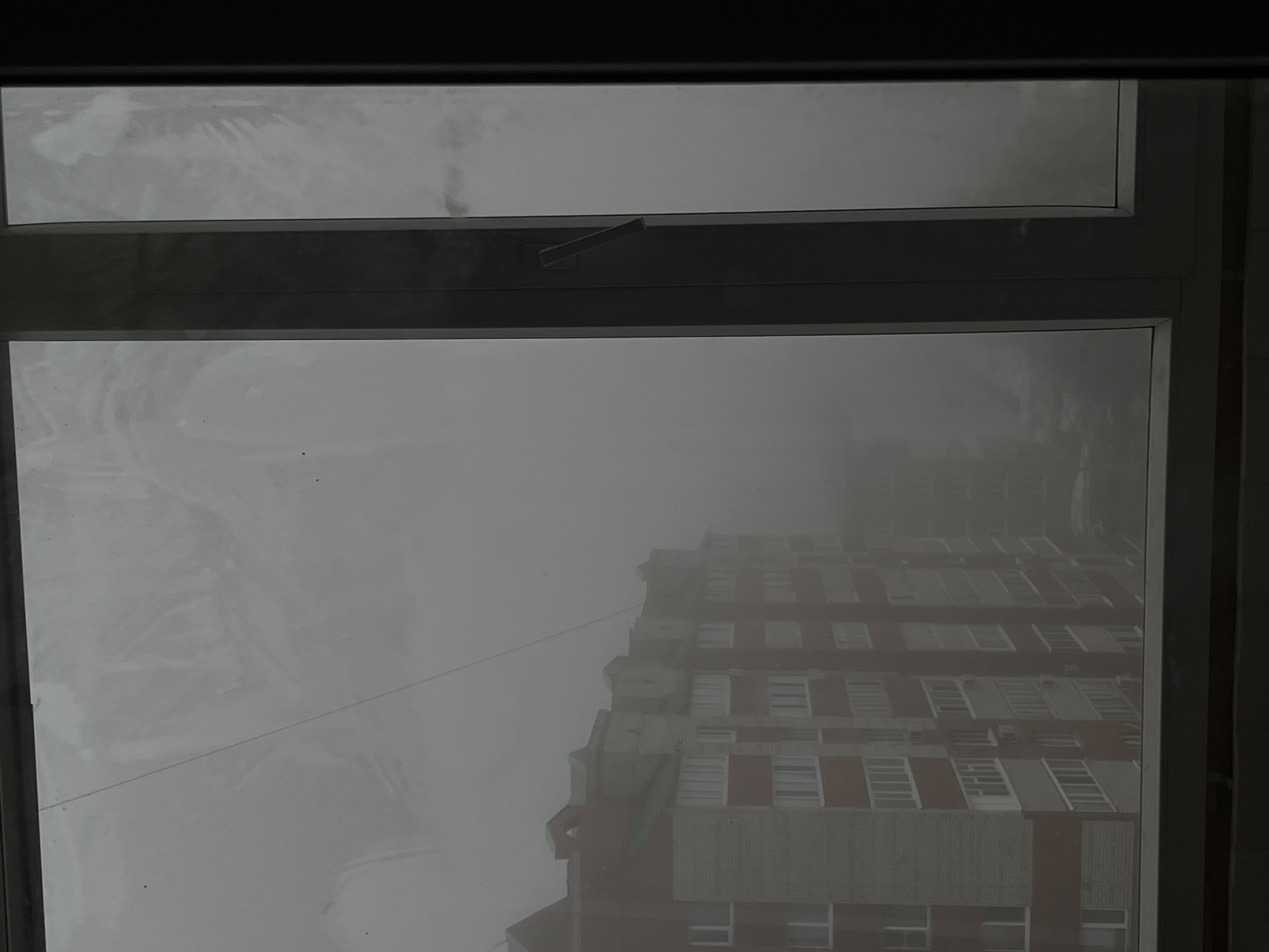 The image is an interior shot of a window with an exterior view dominated by heavy fog. The fog obscures much of the scene outside. A tall building is partially visible through the fog, its facade featuring rows of windows. The building's brick is a muted reddish-brown, contrasting slightly with the overcast sky. The window frame is dark gray. A window handle is positioned on the top window pane. The overall atmosphere is gray and bleak. The location is believed to be in the city of Neftekamsk, Russia.