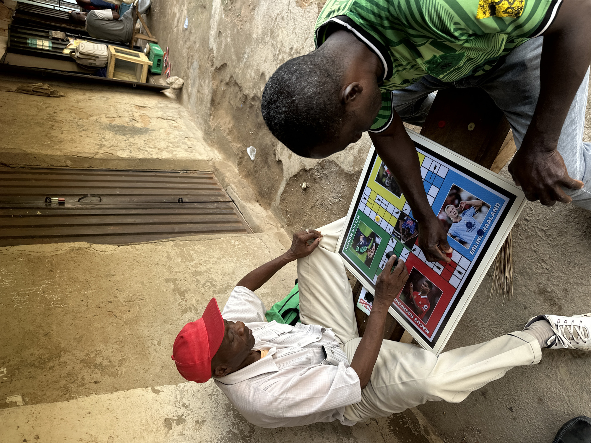 In Narayi, Nigeria, two men are engaged in a board game outdoors. The man on the left, wearing a red cap and a light-colored striped shirt, is seated on a wooden bench. He is focused on the board, which features images of soccer players. The man on the right, wearing a green and black striped shirt, is also seated and participating in the game. The board game appears to be placed on a wooden surface. The background consists of a textured, weathered wall and a metal corrugated door. The ground is rough and appears to be dirt or concrete. There are other objects visible in the background, including bags and crates, suggesting a commercial or roadside setting.