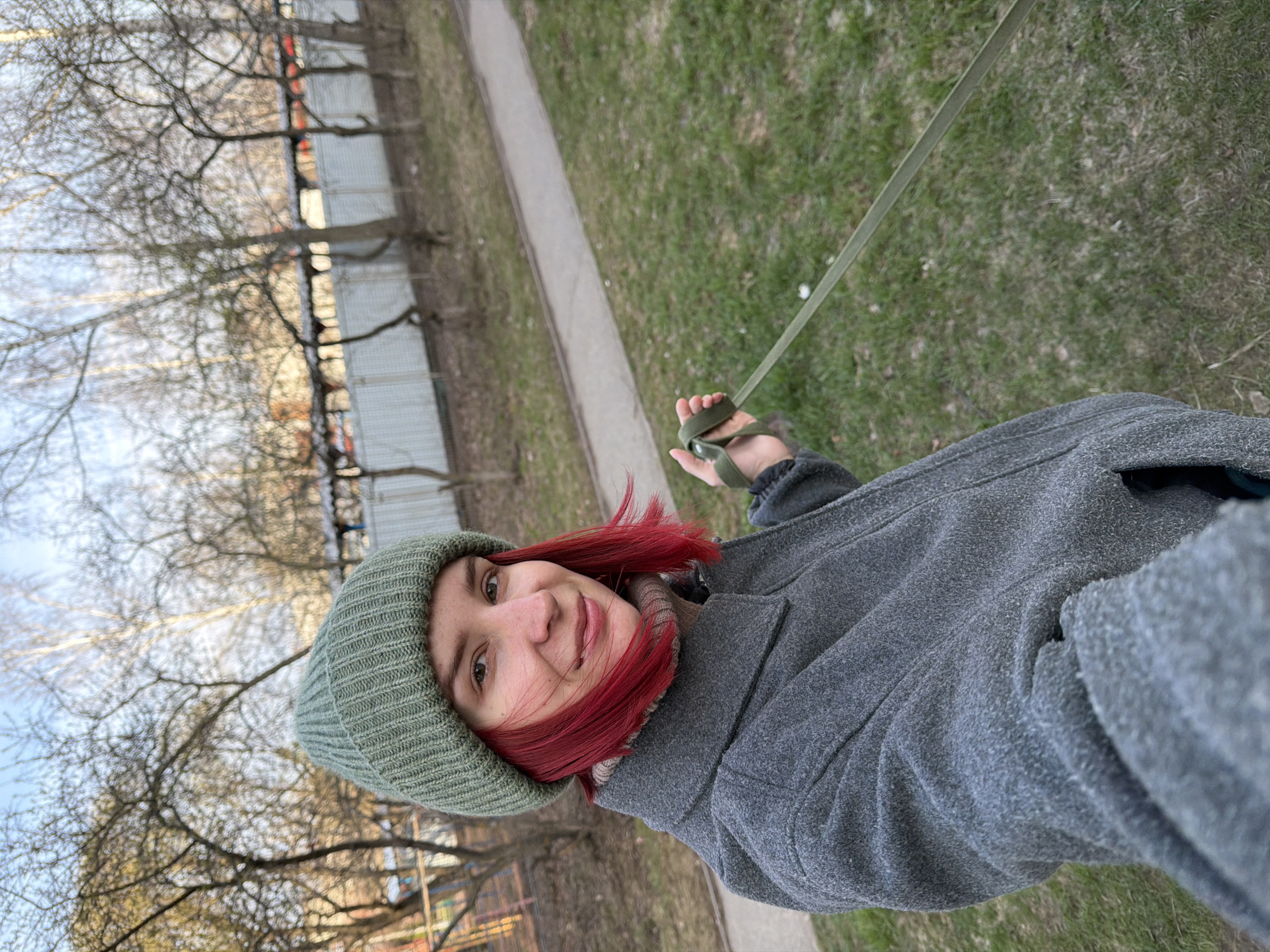 A woman with red hair and a green beanie is holding a leash while standing outside in Chaikovsky, Russia. She is wearing a gray coat. The background includes a sidewalk, grass, trees, and a building.