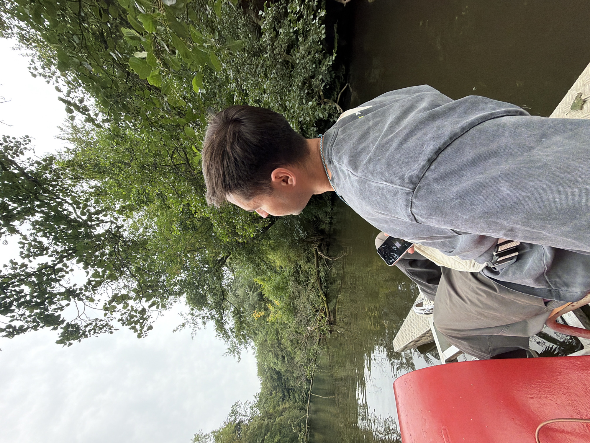 A person is seated in a red pedal boat on a narrow waterway, likely in Aalsmeer, Netherlands, given the lush green trees and calm water. The individual is viewed from behind and to the right, wearing a grey oversized sweatshirt and dark pants. They are holding a mobile phone in their right hand and looking off into the distance at the dense foliage lining the water's edge. The water appears dark and reflects the overcast sky and surrounding trees. The boat has a metal platform in front of the person.