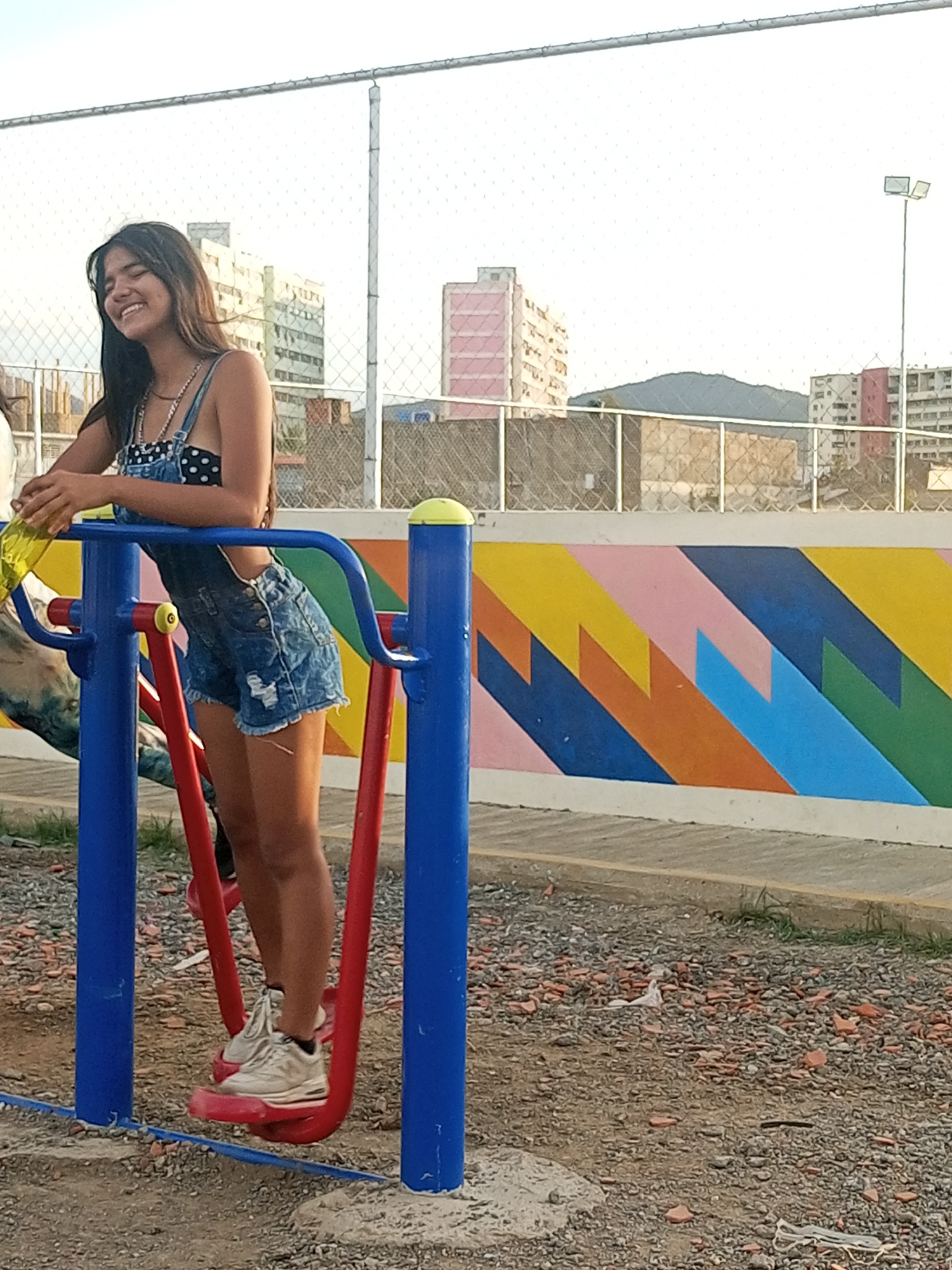 The image features a young woman smiling broadly while engaged with an outdoor exercise machine, likely an air walker or elliptical, in a public park or community space in Juan Griego, Venezuela. She has long, dark hair and is dressed in a black and white polka dot top layered under blue denim overall shorts, accessorized with several necklaces, and wearing white sneakers. She holds onto the blue handles of the red and blue exercise equipment, with her feet on the platforms, and a yellow bottle is visible near her left hand.
The setting includes a ground covered in gravel and small stones. Behind the woman, a vibrant mural displays a geometric pattern of colorful chevrons and zigzags in shades of yellow, orange, blue, pink, and green on a low wall. Above this, a chain-link fence separates the area from the background. Beyond the fence, various multi-story buildings, some with prominent red or pink facades, are visible, along with a distant mountain range under a bright, clear sky, suggesting a sunny day, possibly late afternoon. A lamppost stands on the far right. The woman's joyful expression conveys a sense of happiness and ease. No discernible text is visible in the image.