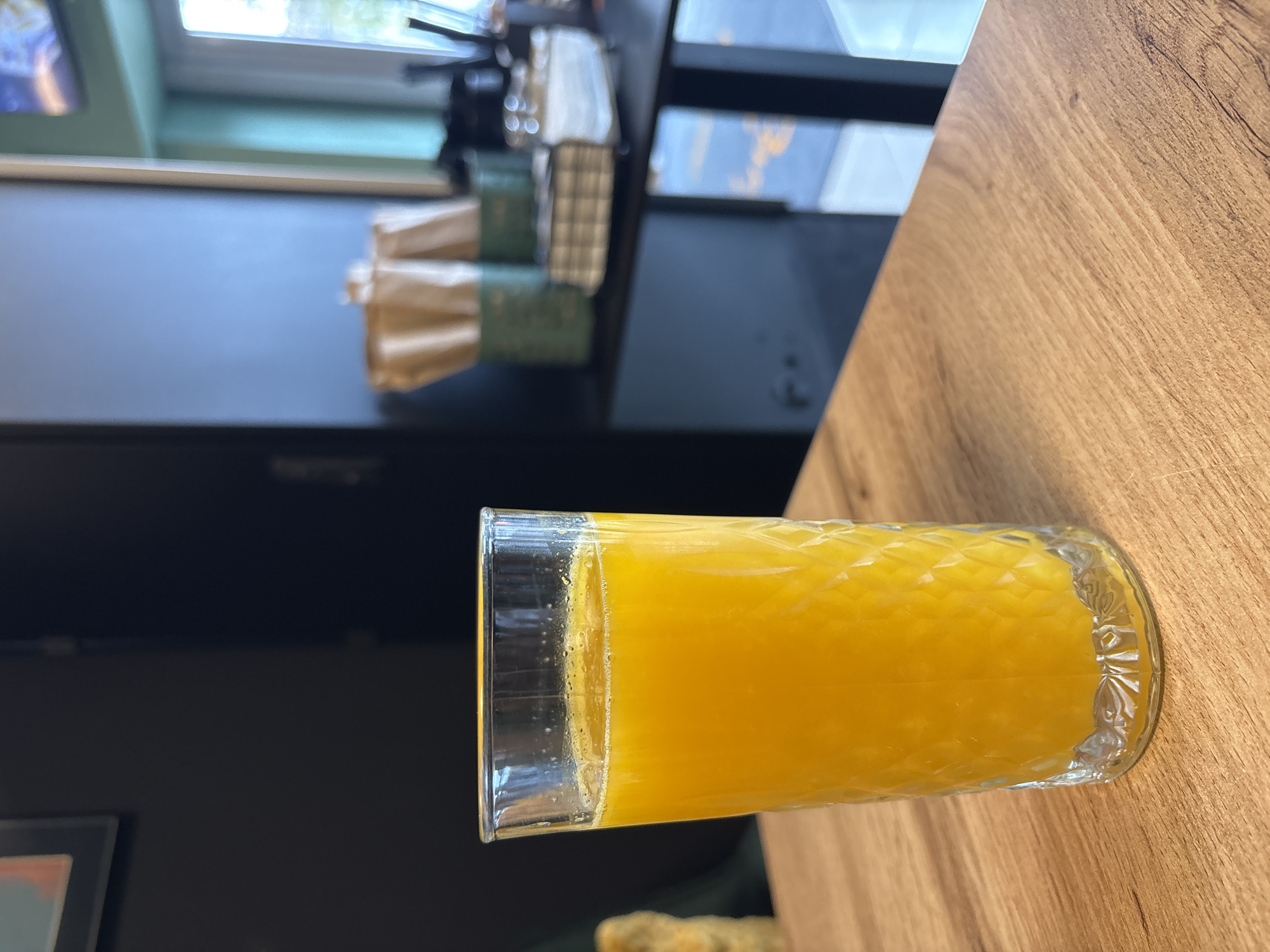 A clear glass filled with bright orange juice stands prominently on a light brown wooden table, which displays a distinct grain pattern. The glass has a textured, possibly diamond-cut, design on its lower half, and small bubbles cling to its inner surface. The background is softly blurred, indicating a shallow depth of field. To the left, a dark surface, possibly a wall or counter, is visible. Further back, a window lets in bright daylight, with blurry green foliage visible outside, suggesting an outdoor view. On a shelf or counter in the mid-ground, several small, neatly stacked packages, appearing to be wrapped in a combination of green/teal material and brown paper, are visible. The setting appears to be indoors, likely a cafe or restaurant, during daytime. There are no people visible, and no discernible text. The scene is situated in Veliko Tarnovo, Bulgaria.