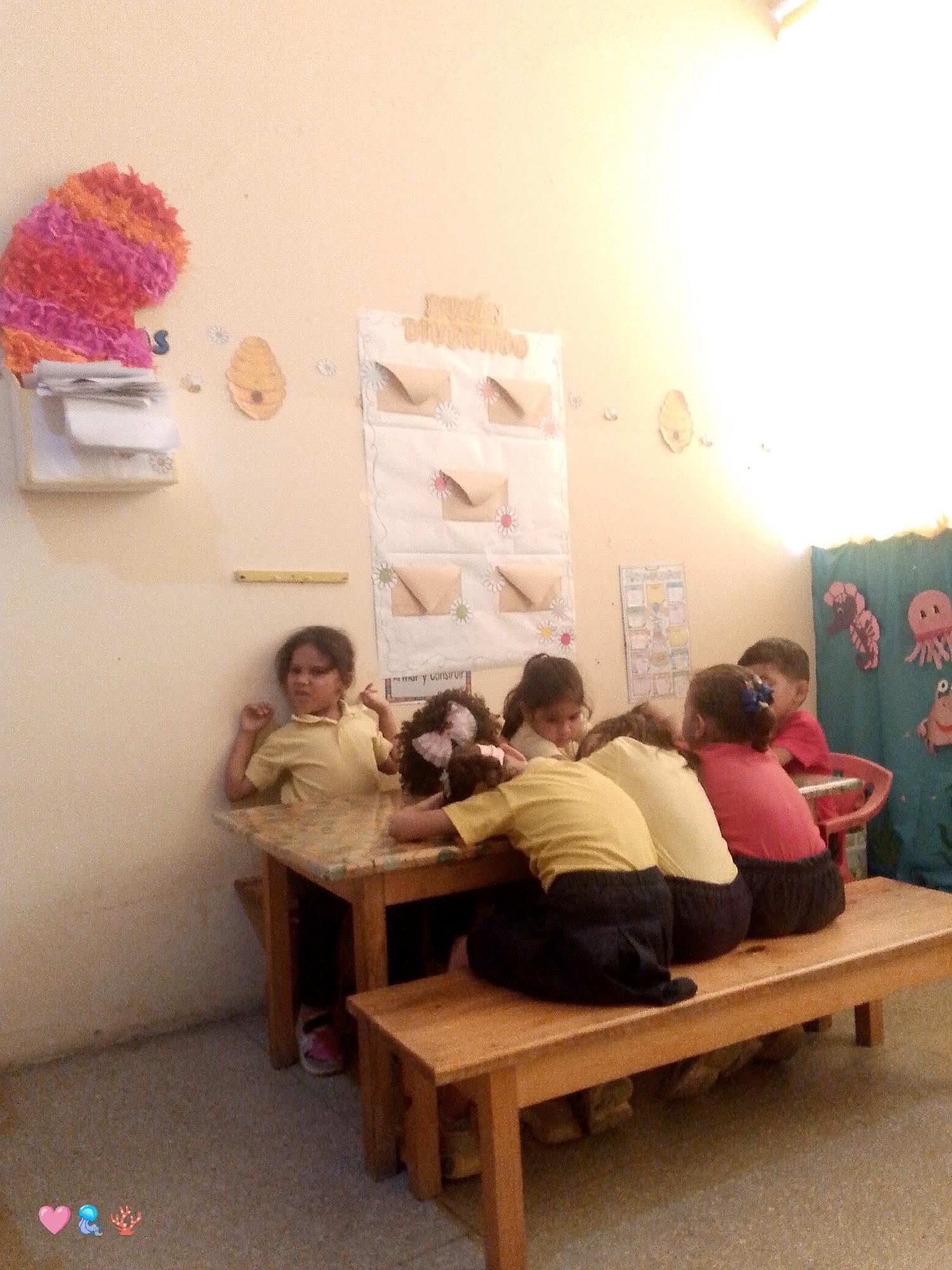 The image captures an indoor scene, likely a classroom or daycare center in Santa Ana, Venezuela, during daytime. Six young children are gathered around a small wooden table. One child, wearing a yellow collared shirt, sits at the left end of the table, looking towards the camera with a neutral to slightly displeased expression. Another child with a pink bow in her hair, also in a yellow shirt, has her head resting on the table, seemingly tired or resting. Adjacent to her, another child in a light-colored shirt also has their head down. To their right, a child in a red shirt and black bottoms is seated on the wooden bench, facing right, with another child in red visible next to them. A sixth child is partially visible on the far right, seated on the bench.
The room is decorated with various items typical of a children's learning environment. On the left wall, there's a vibrant, multicolored paper decoration (possibly a cut-up piñata or abstract art) above a white wall shelf holding papers. Further right on the wall are two yellow beehive cutouts and several small white flower decorations. A prominent white fabric wall organizer features multiple small brown paper envelopes and flower motifs. Visible text on this organizer includes "DIVIERTO" at the top and partially "amor y convivir" (love and coexist/live together) further down. A yellow ruler-like object is also affixed to the wall. To the far right, a blue fabric curtain adorned with marine life cutouts, such as a lobster/crab and an octopus, adds to the cheerful decor. The floor is a speckled, light-colored surface. Bright light, possibly from a window or overhead fixture, illuminates the scene, creating a glare in the upper right.
