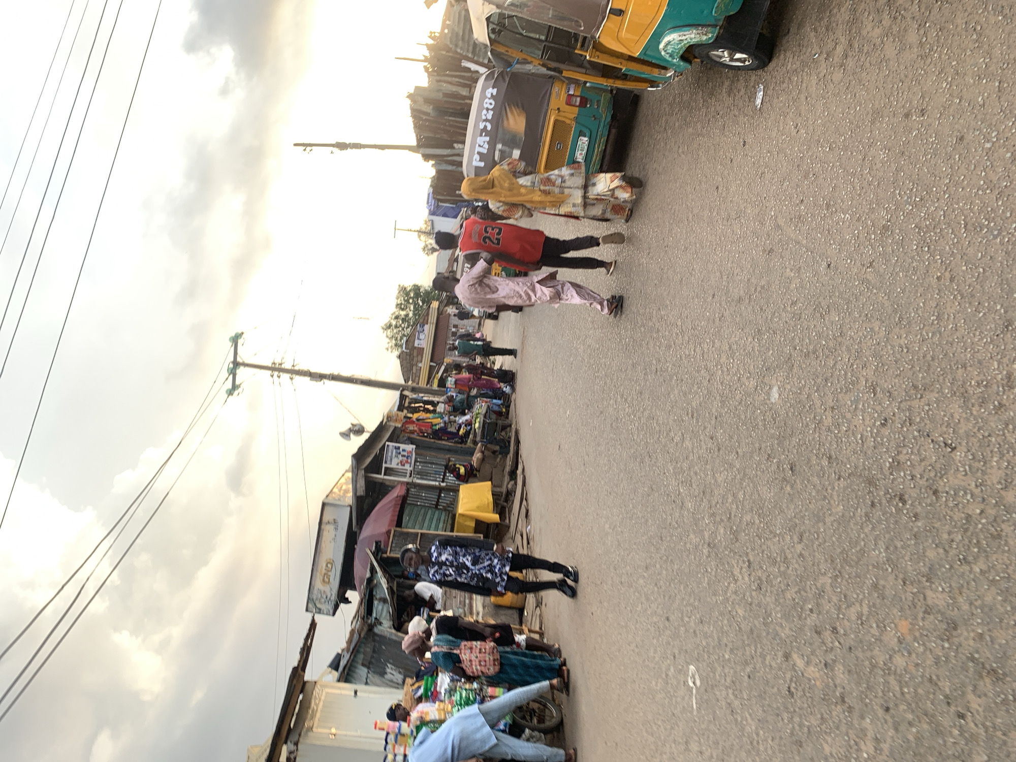 The image shows a street scene in Gura Top, Nigeria. The street is paved and busy with pedestrians and motorized vehicles. A yellow and green tuk-tuk is parked at the top of the frame. Several people are walking along the street, including two men walking together with a man wearing a red basketball jersey with the number "23" on the back. Other people are standing or walking near shops and stalls lining the side of the road. Some shops appear to be permanent structures with corrugated metal roofs. Power lines and poles are visible along the left side of the image. The sky is overcast with diffused sunlight.