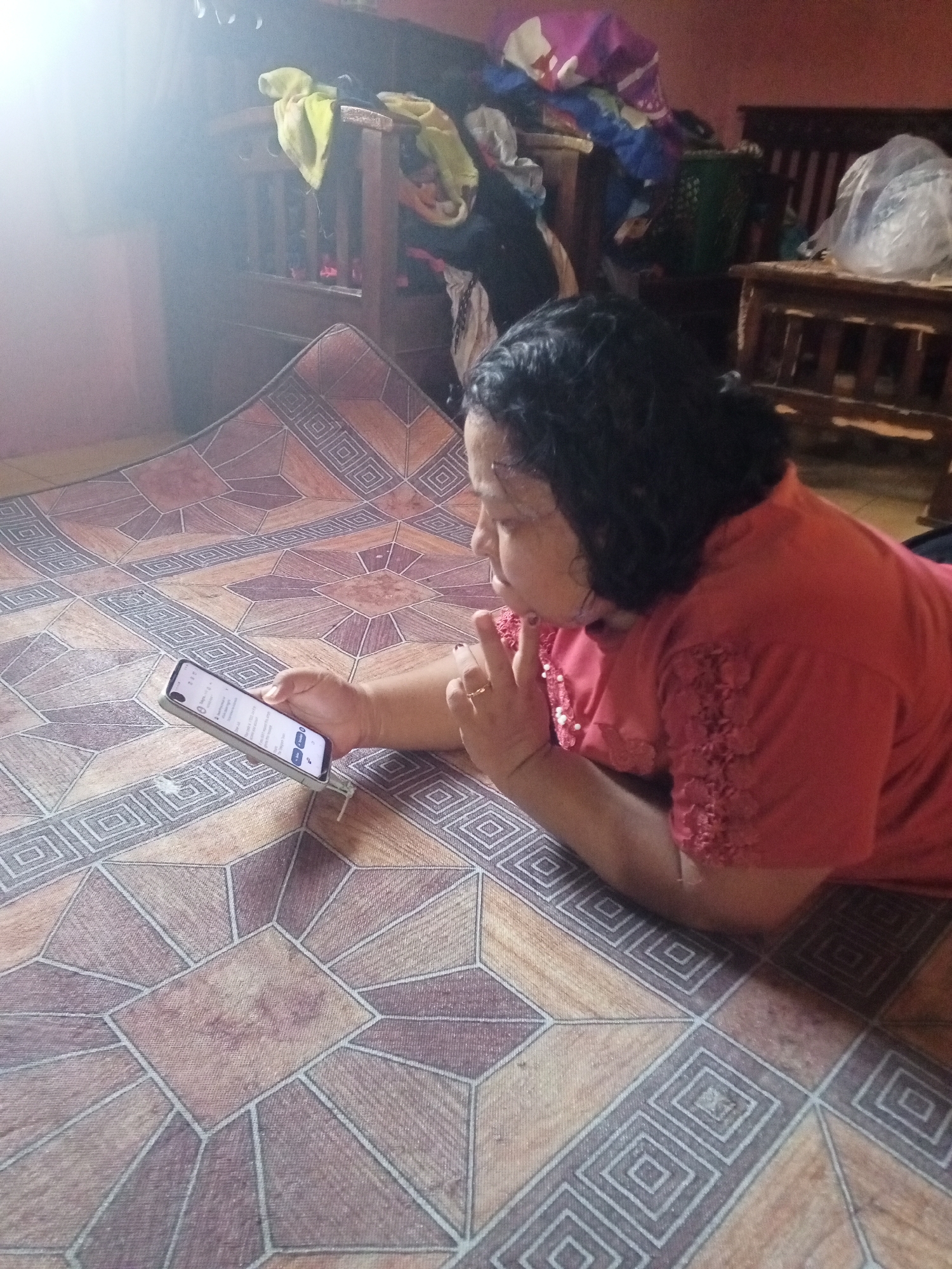 A woman is lying on her stomach on a patterned floor, holding a smartphone. She is wearing a red shirt with lace detailing on the sleeves. Her hair is dark and curly. The floor has a geometric pattern with diamond and square shapes. In the background, there is furniture and some items that appear to be clothes or fabric. The lighting suggests an indoor environment, possibly a home in Tanah Gara Hulu, Indonesia, with light entering from the left.