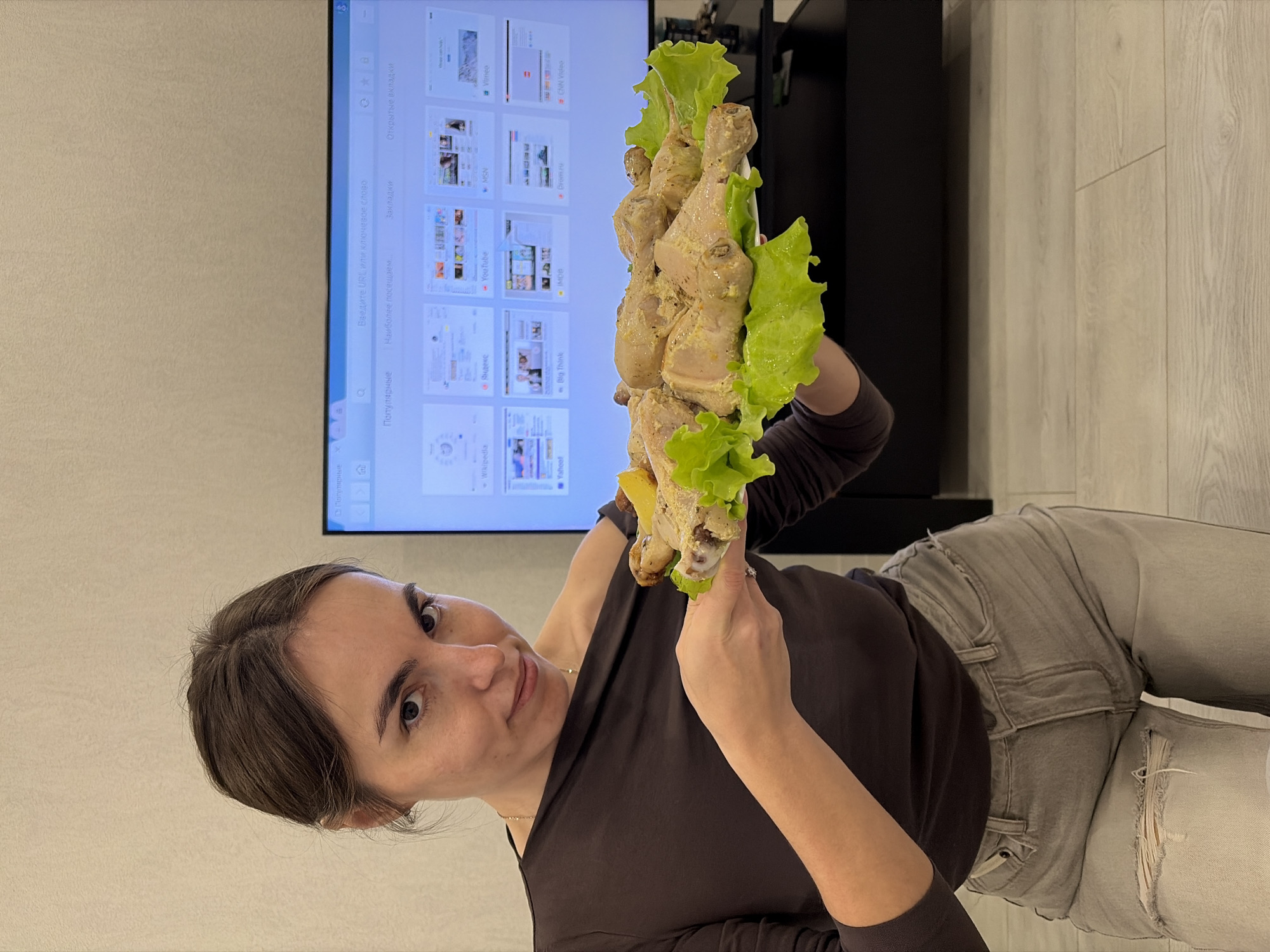 A woman is holding cooked chicken legs garnished with lettuce leaves. She is indoors, likely in a home. Behind her is a wall-mounted television displaying internet browser tabs in Russian, including tabs for "Wikipedia", "Yandex", "YouTube", "Vimeo" and "CNN Video". The woman is wearing a dark brown, long-sleeved shirt and light gray jeans with a ripped area on the thigh. The location in the city of Sarapul, Russia cannot be verified based solely on the image.