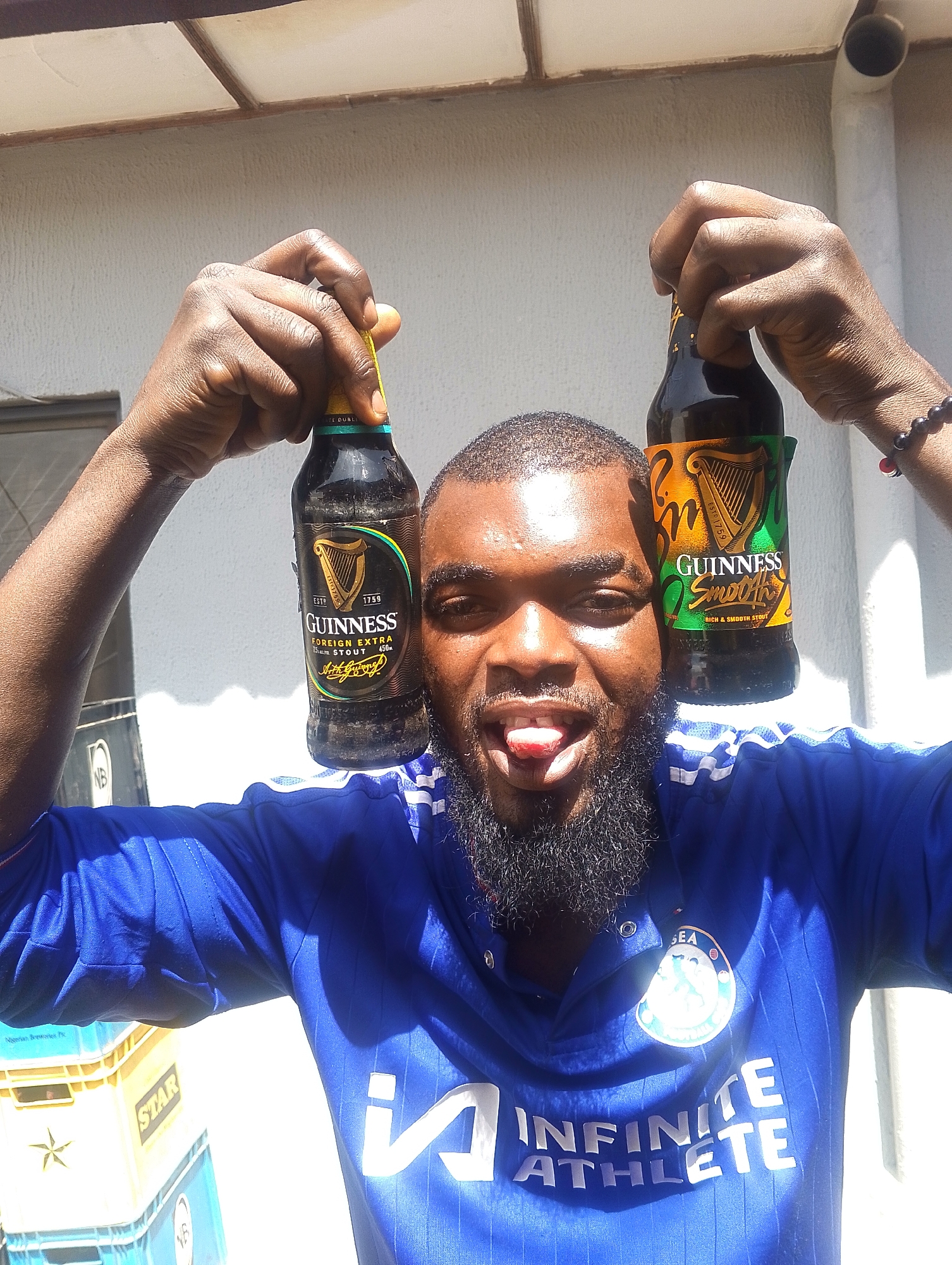 The image shows a Black male in his late 20s or early 30s, with a full beard and dark skin, holding two bottles of Guinness beer, one in each hand. He is wearing a blue jersey with white stripes on the sleeves and a logo on the chest. The man is sticking his tongue out and looking at the camera with a playful expression. Behind him, there are stacks of plastic crates, some of which appear to be for "Star" beer. The background suggests an outdoor setting, possibly a backyard or a street in Jos, Nigeria, with a textured wall and part of a roof visible. The lighting indicates it is daytime.